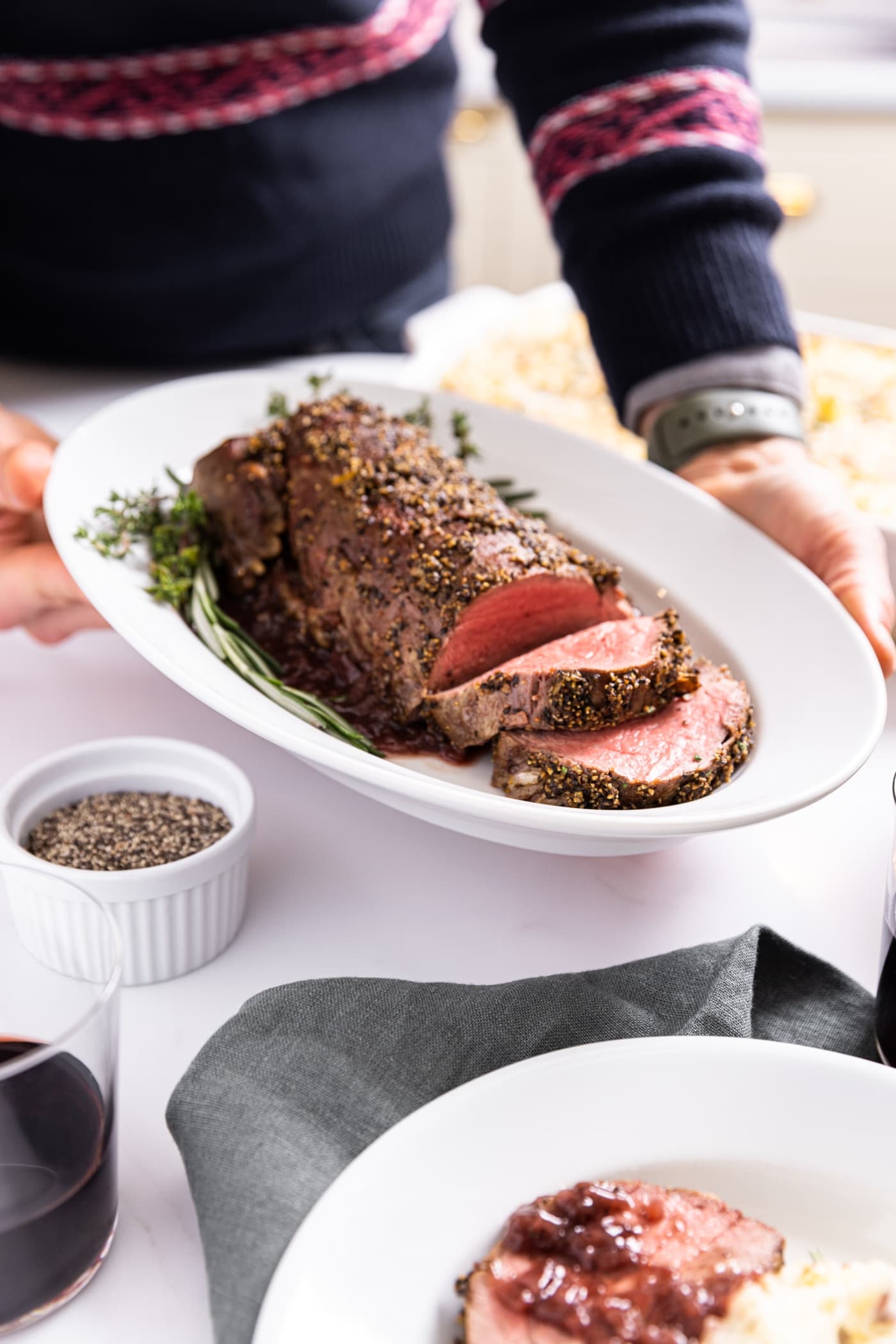 Two hands holding large white platter with peppercorn crusted beef tenderloin with two slices sliced with white plate with mashed potatoes