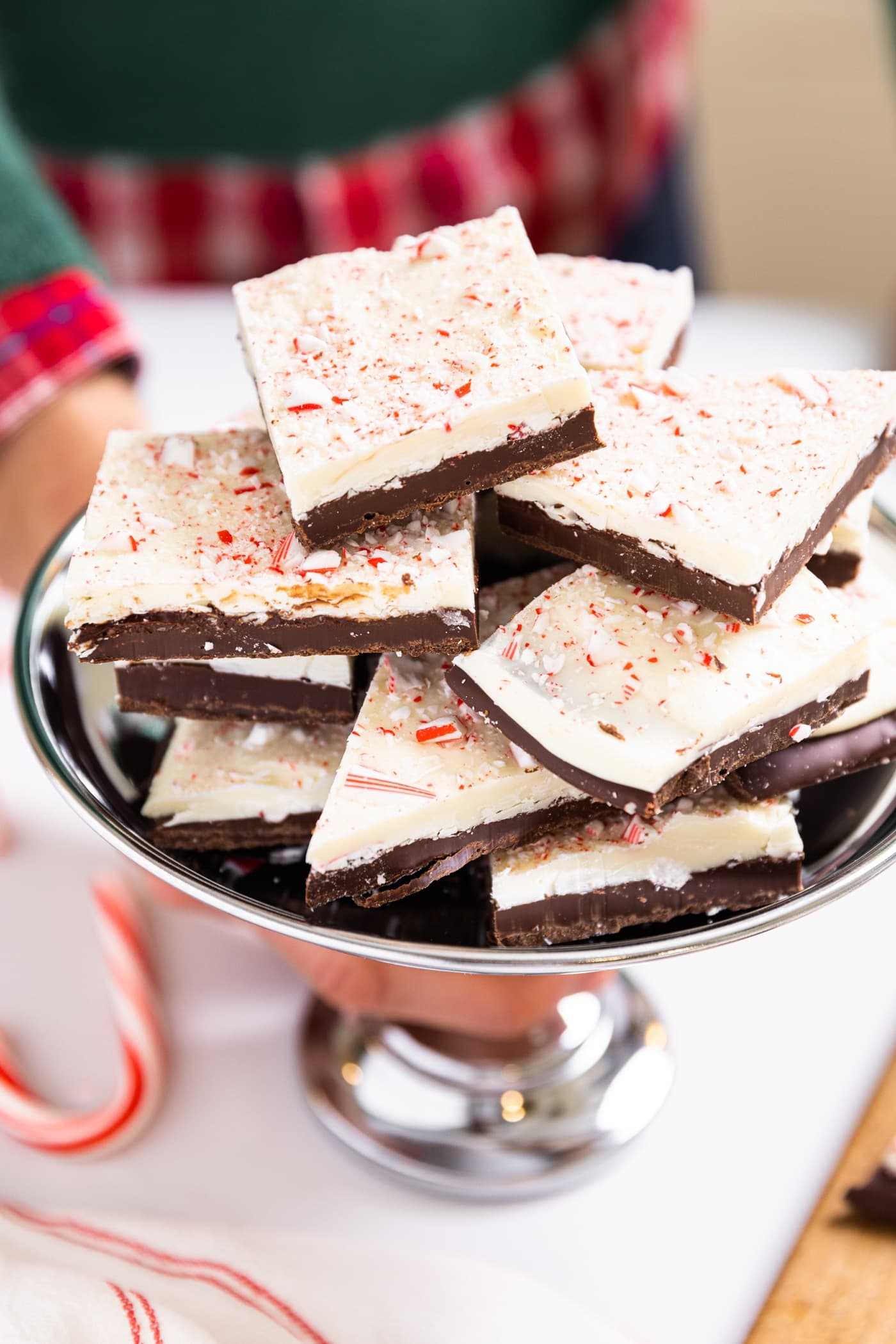 Hand holding silver dish filled with stacks of peppermint bark with chocolate and white chocolate layers and topped with peppermint pieces