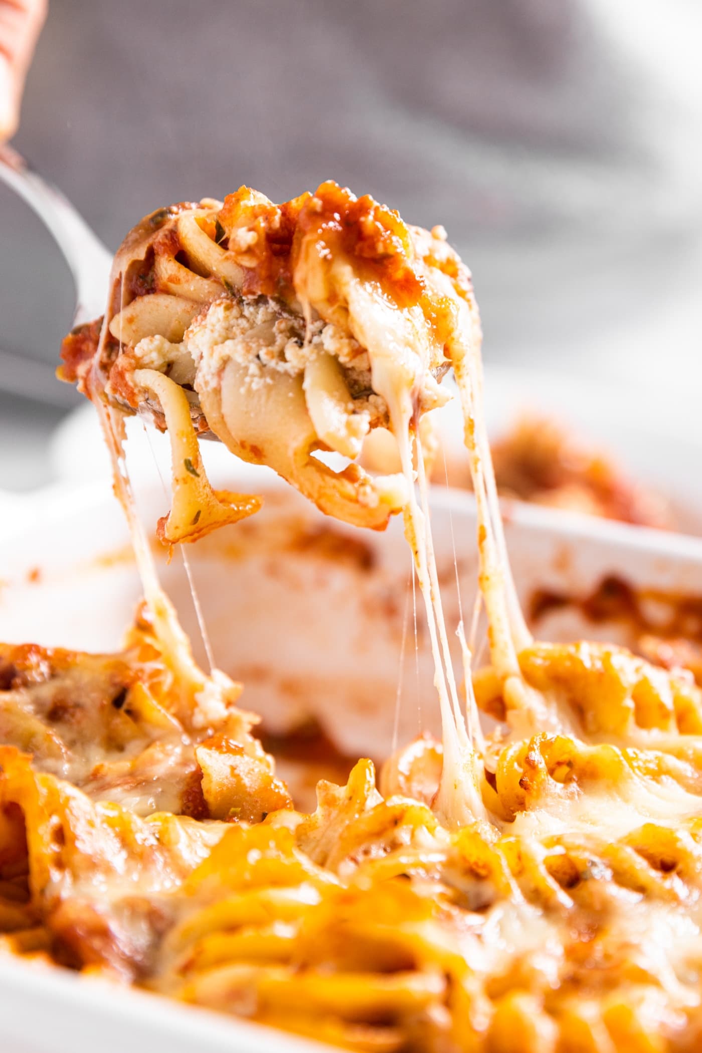 Spoon pulling piece of baked rotini casserole from baking dish with cheese strings clinging to the rest of the casserole
