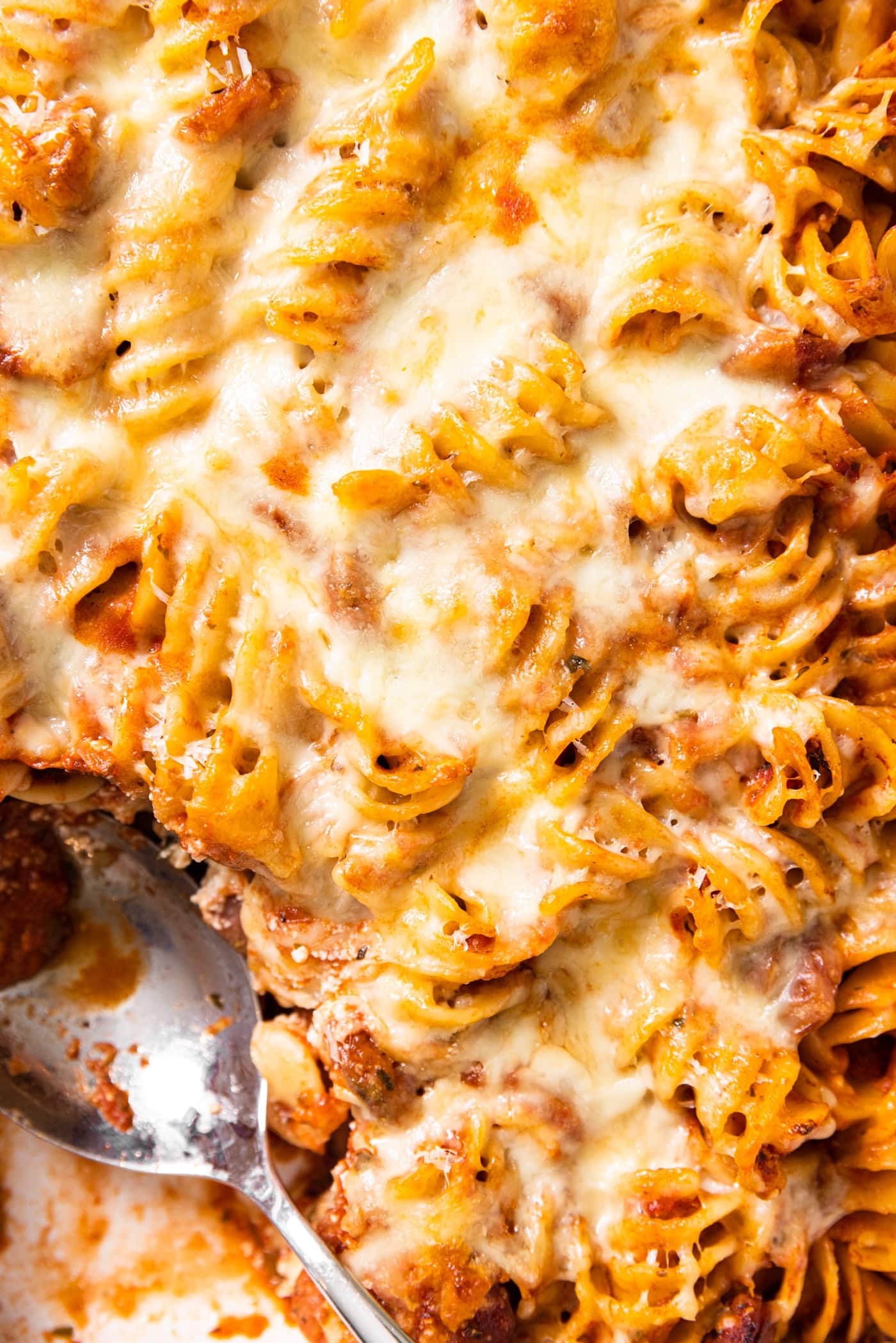 Top down view of baked rotini casserole in baking dish with cheese all over the top with burnt crisp edges showing and spoon sitting in dish