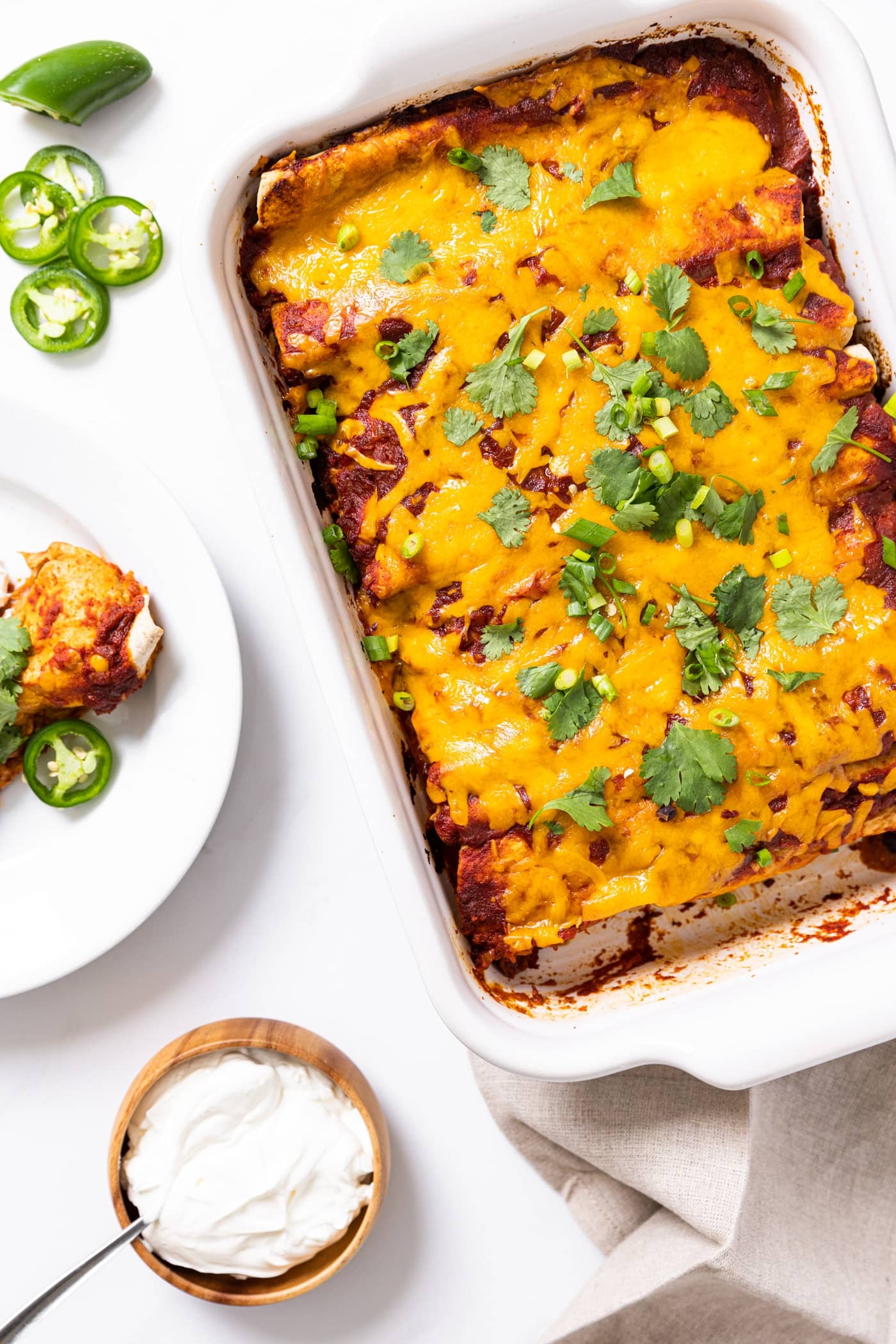 Top down view of white baking dish filled with chicken enchiladas topped with cilantro and plate filled with one slice of enchilada