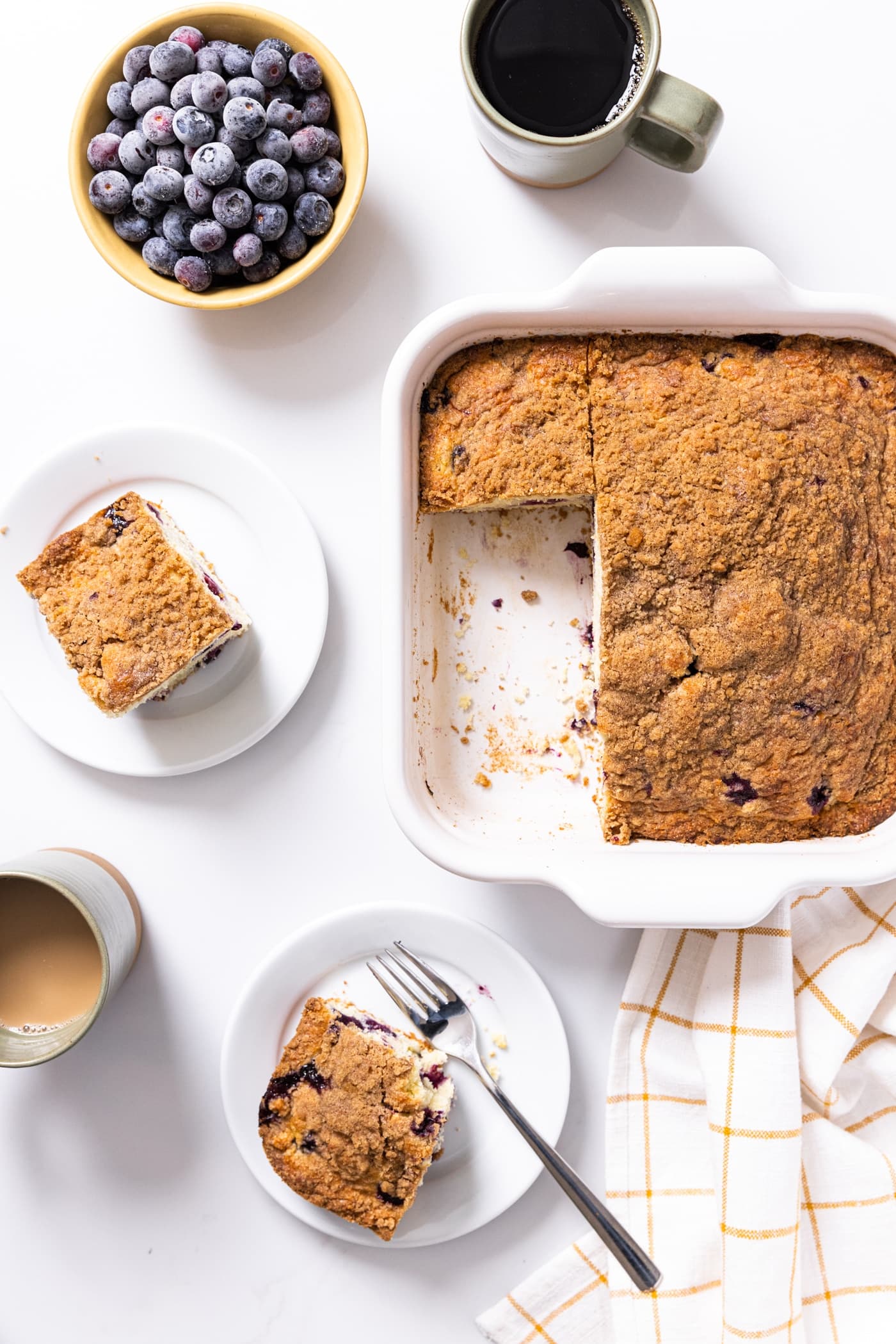Top down view of blueberry coffee cake sitting in white baking dish with slice taken out sitting on white plate along with coffee cups filled with coffee
