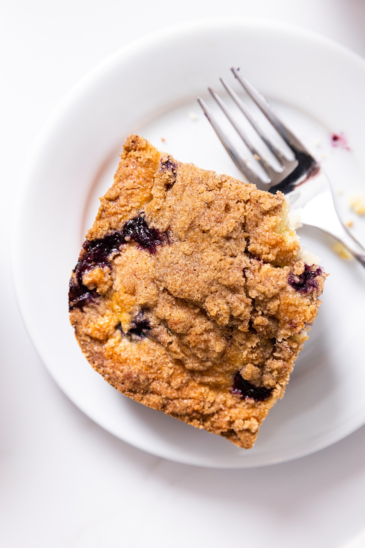 Top down view of piece of blueberry coffee cake with crisp topping sitting on white plate with a fork