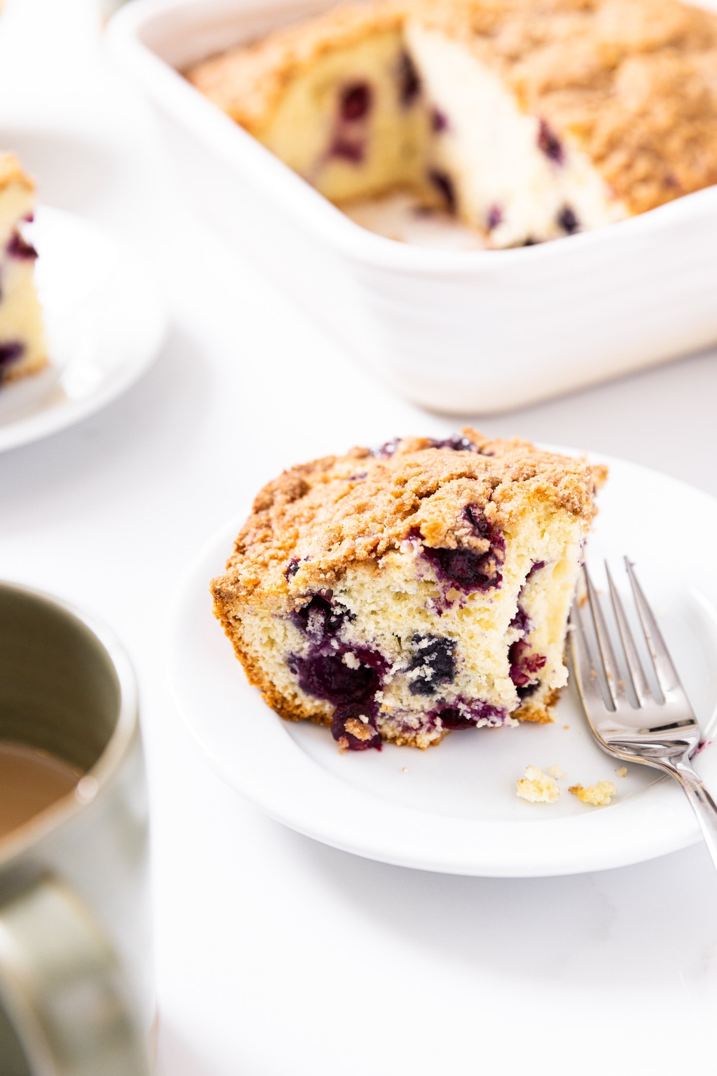 White plate with piece of blueberry coffee cake sitting on white plate with fork and rest of coffee cake in background