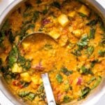 Top down view of metal kettle filled with orange colored lentil soup with ladle sitting in middle of soup all on white countertop