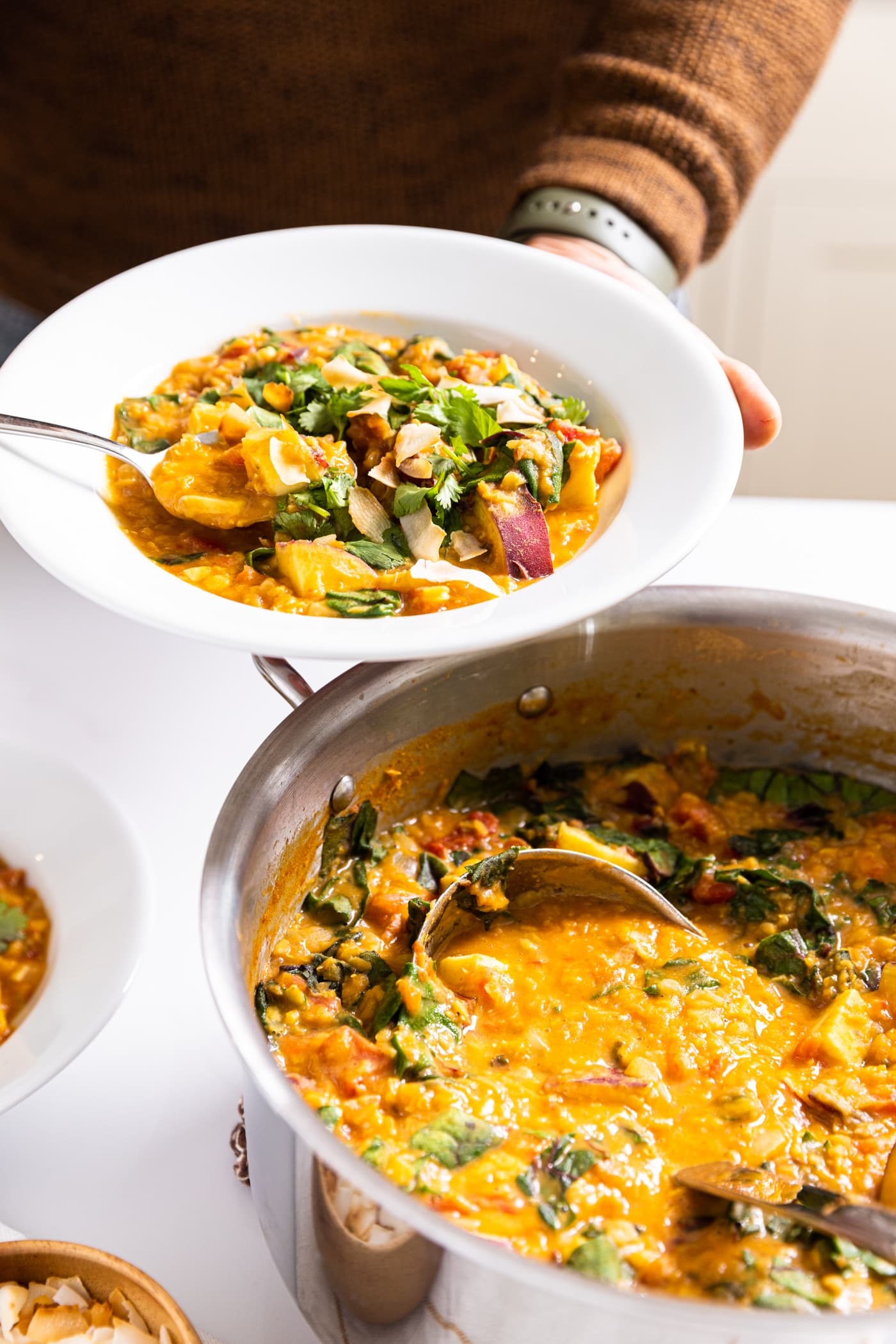 Two hands holding white bowl filled with orange colored creamy curry lentil soup with spoon scooping up piece of sweet potato with rest of soup in kettle sitting on white countertop below