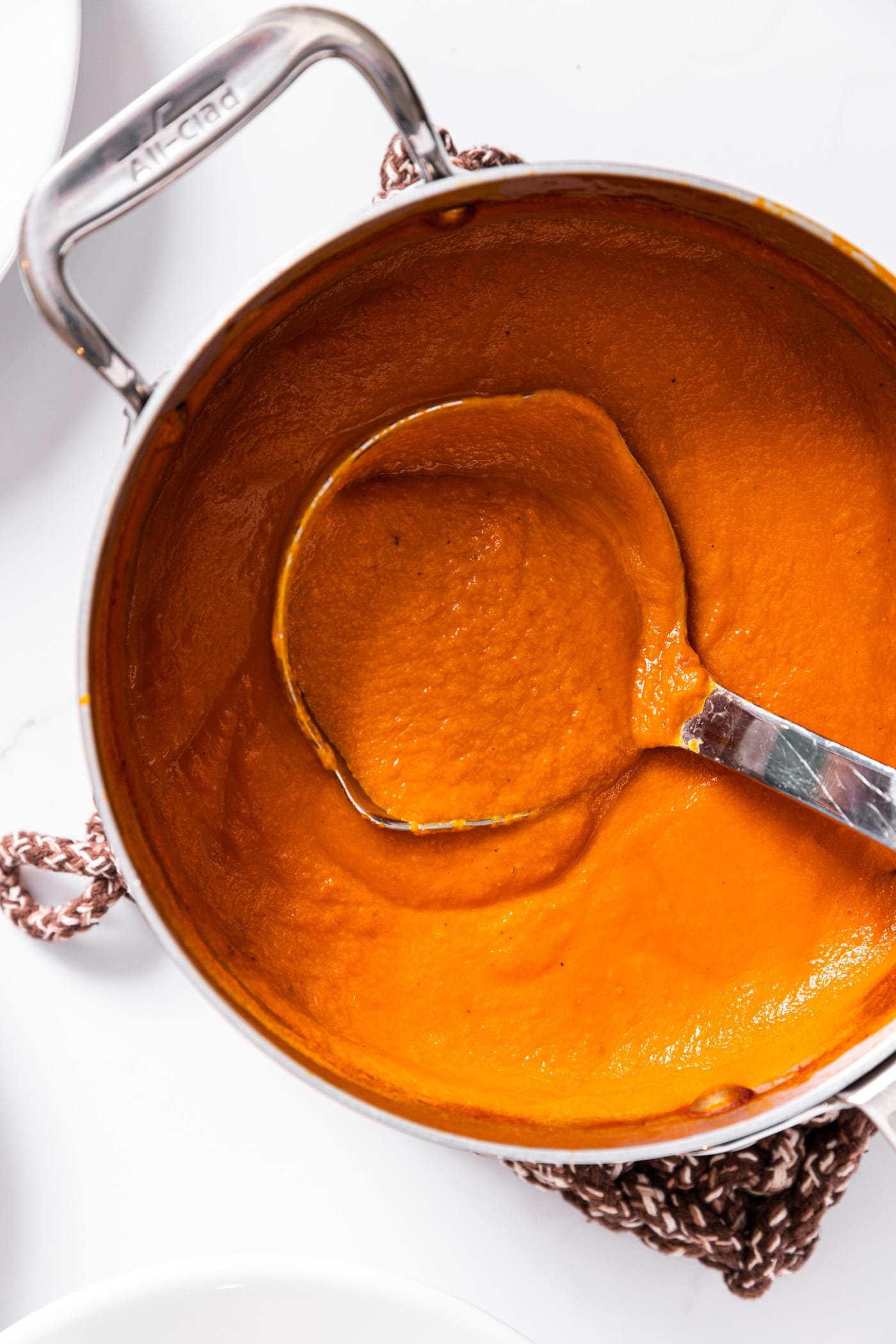 Top down view of metal pot filled with creamy tomato soup with ladle inside all on white countertop