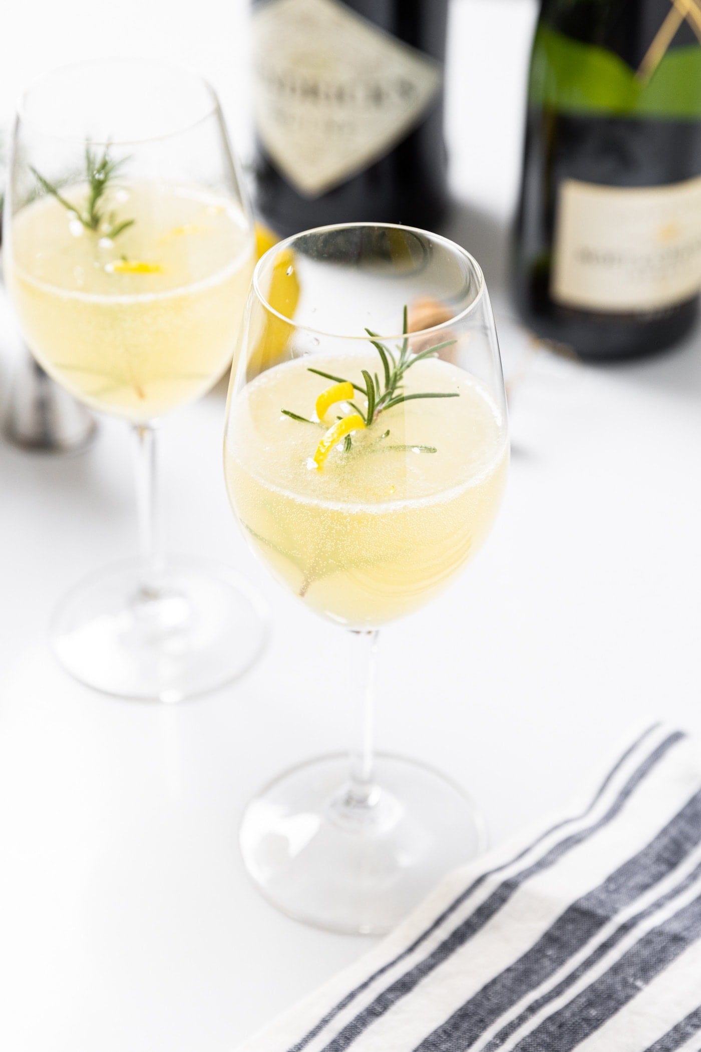 Two wine glasses sitting on white countertop with French 75 cocktail in each one with sprig of rosemary and lemon peel twist with bottles of alcohol in background