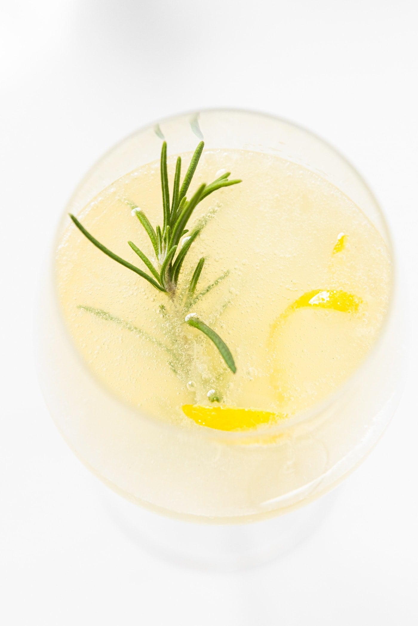 Top down view of wine glass filled with French 75 cocktail with sprig of rosemary and lemon zest peel all on white countertop