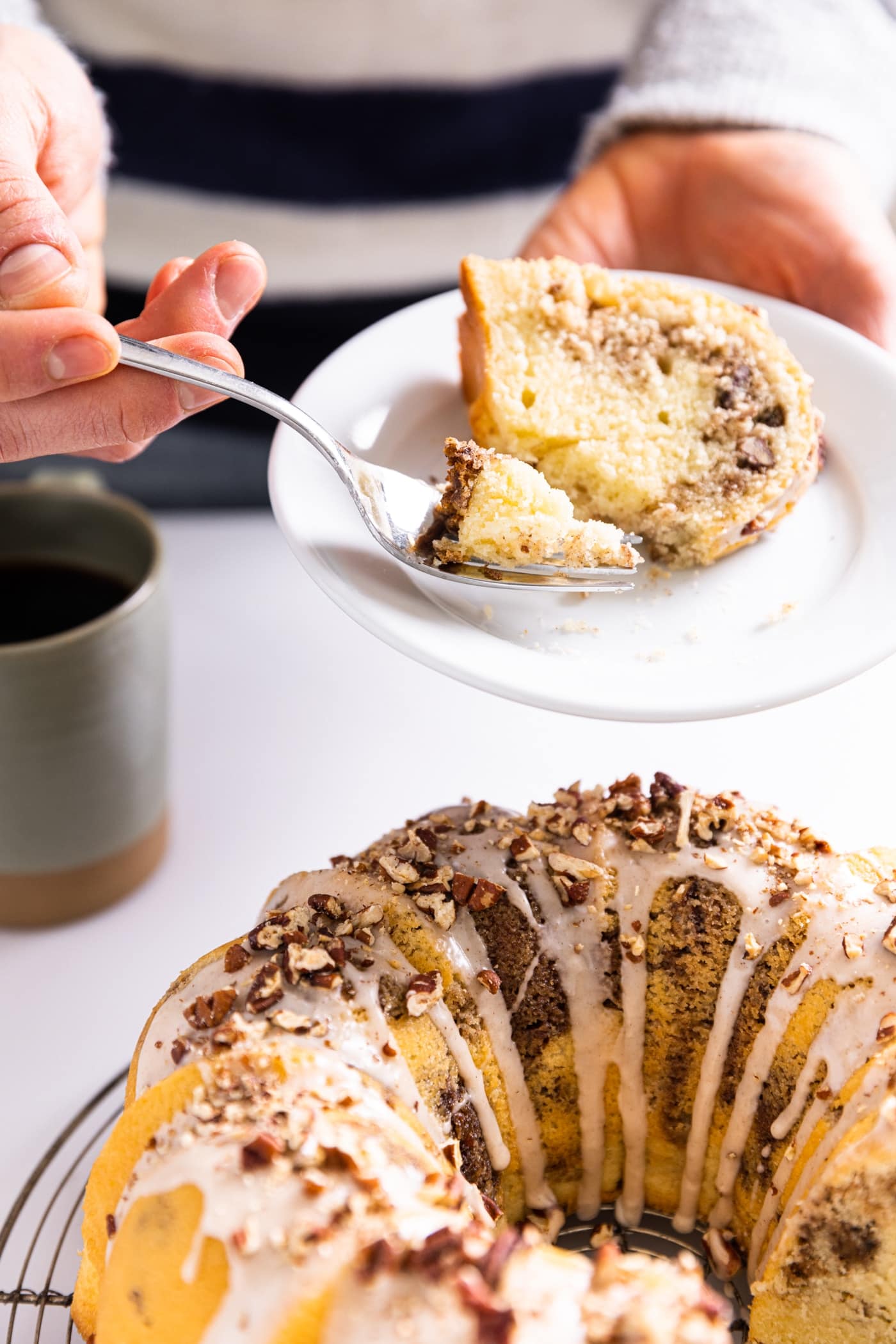 Hand holding white plate with slice of cinnamon sugar coffee cake topped with white icing and chopped pecans with rest of coffee cake sitting on counter below