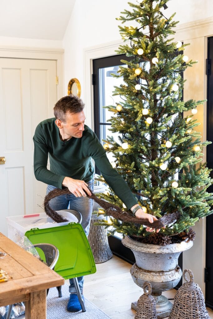 Man taking grapevine garland from partially-empty Christmas tree sitting in an urn