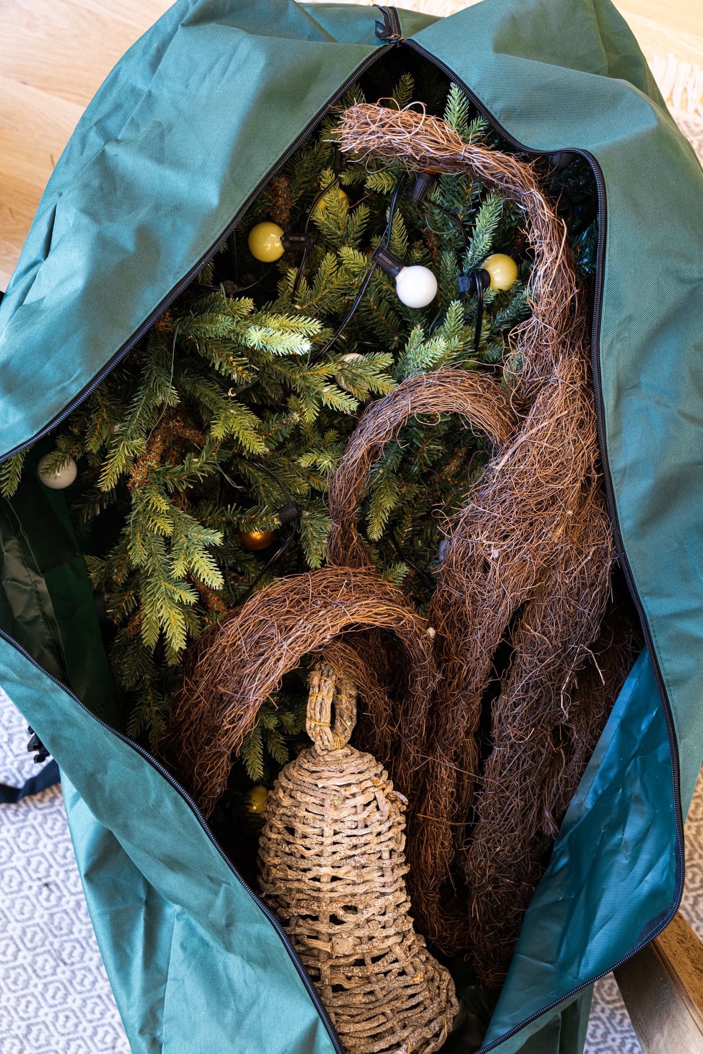 Large green tree bag sitting on floor with part of fake branches from tree sitting inside along with a straw bell and grapevine garland