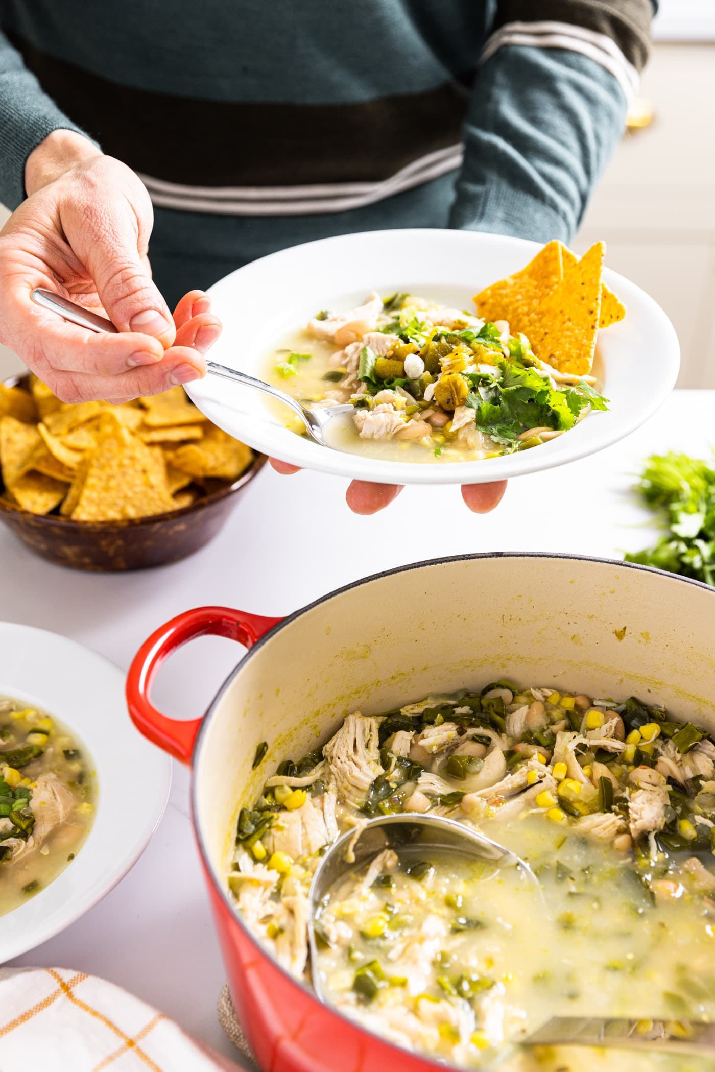 Hand holding white bowl filled with white bean chicken chili scooped from large red Dutch oven below