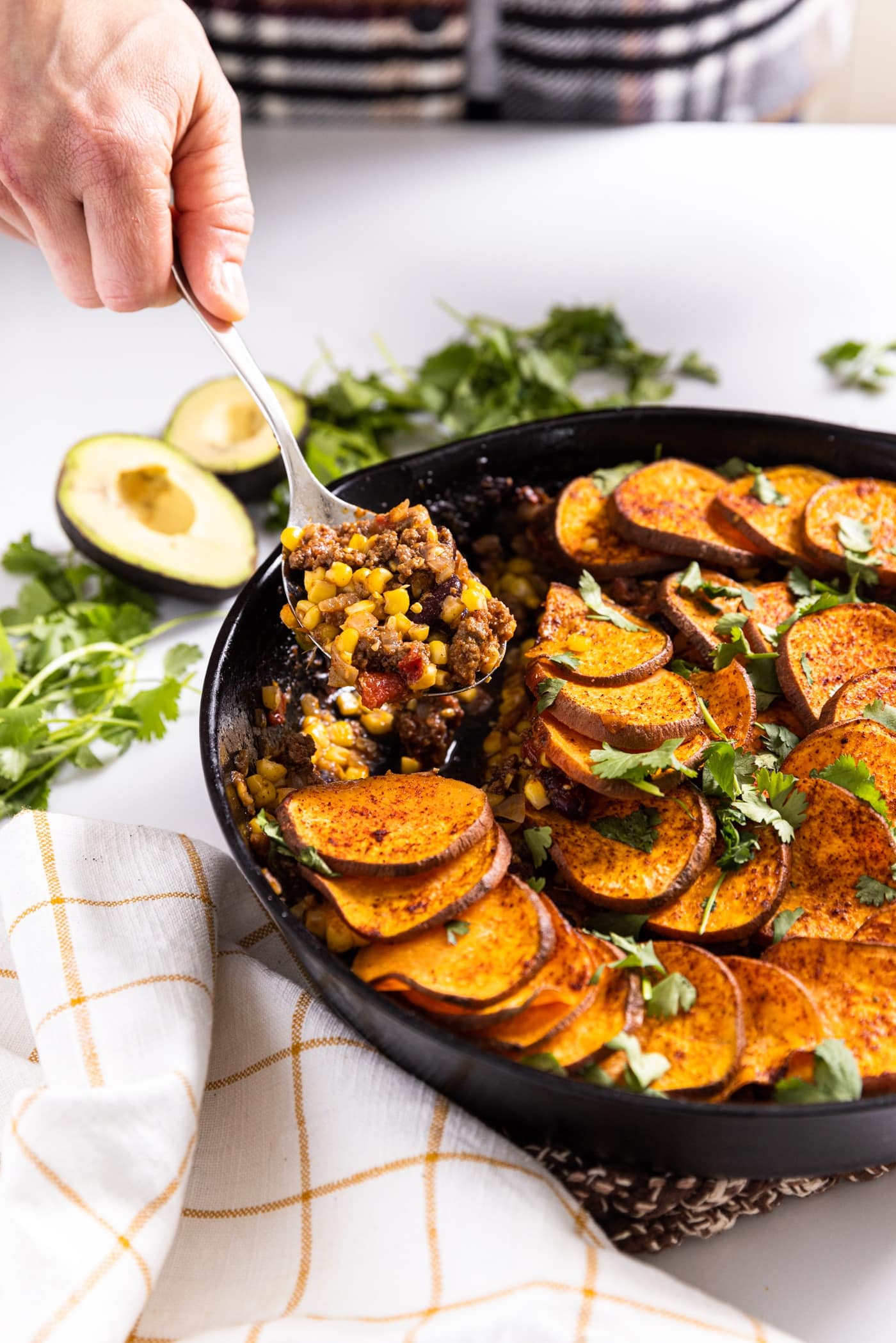 Hand holding spoon scooping out a serving of taco bake topped with slices of sweet potato all on white countertop