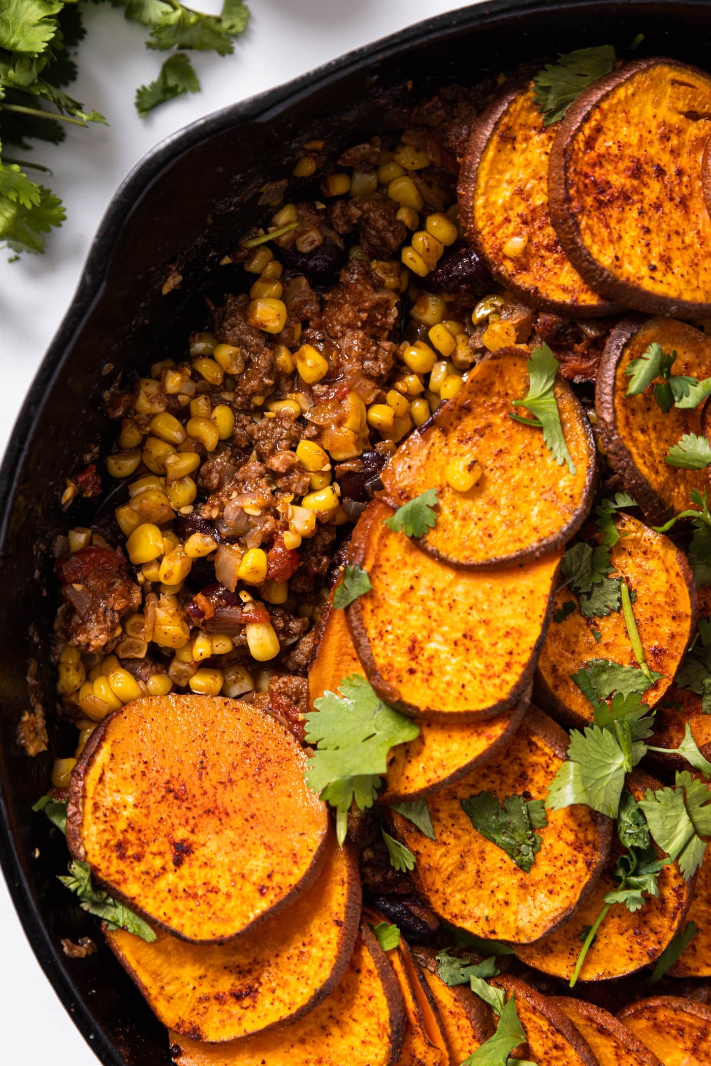 Top down view of taco bake sitting in black skillet with sweet potatoes shingled on top with cilantro on top
