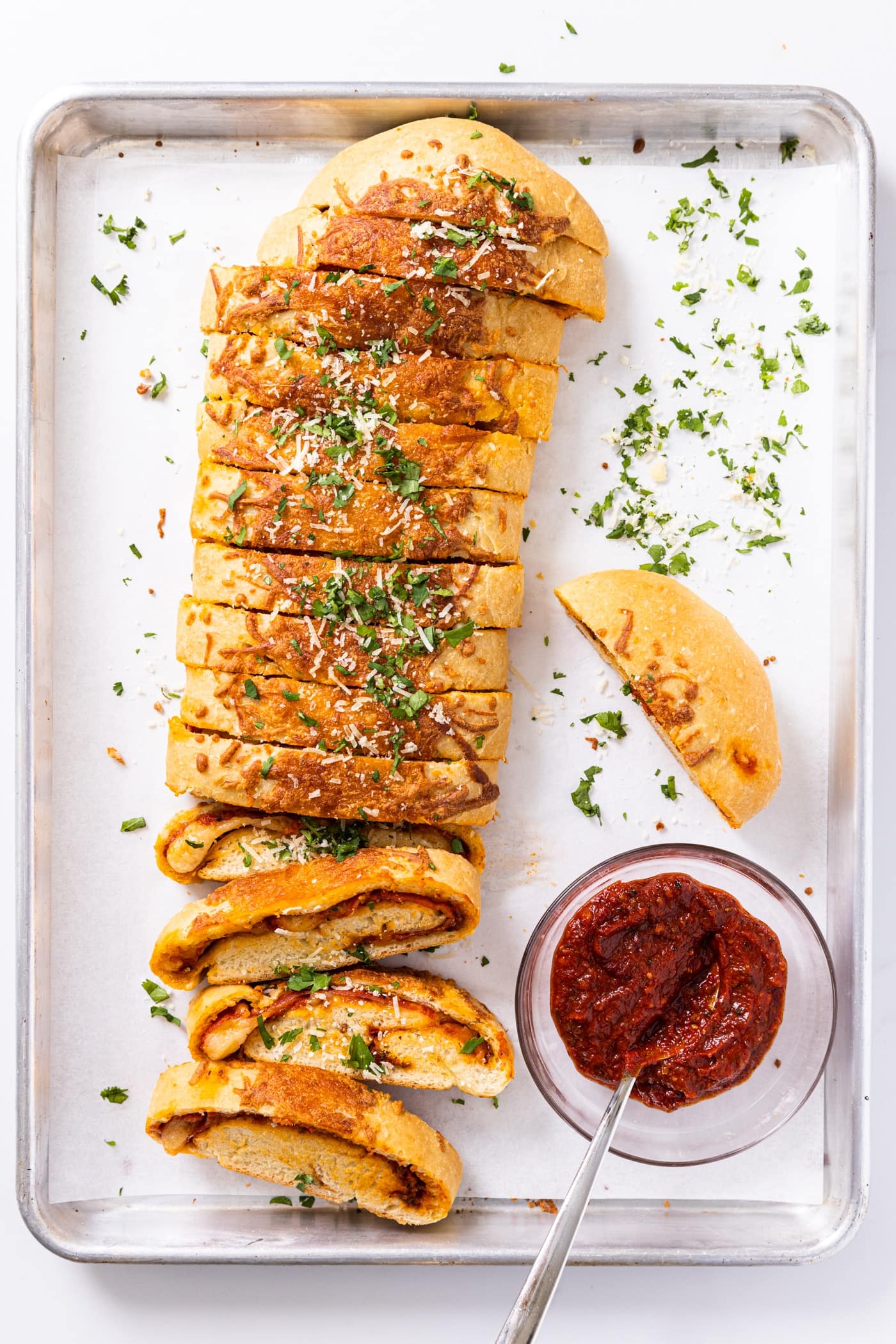 Top down view of metal baking sheet with long sliced stromboli sitting on white parchment paper with small bowl of red tomato sauce