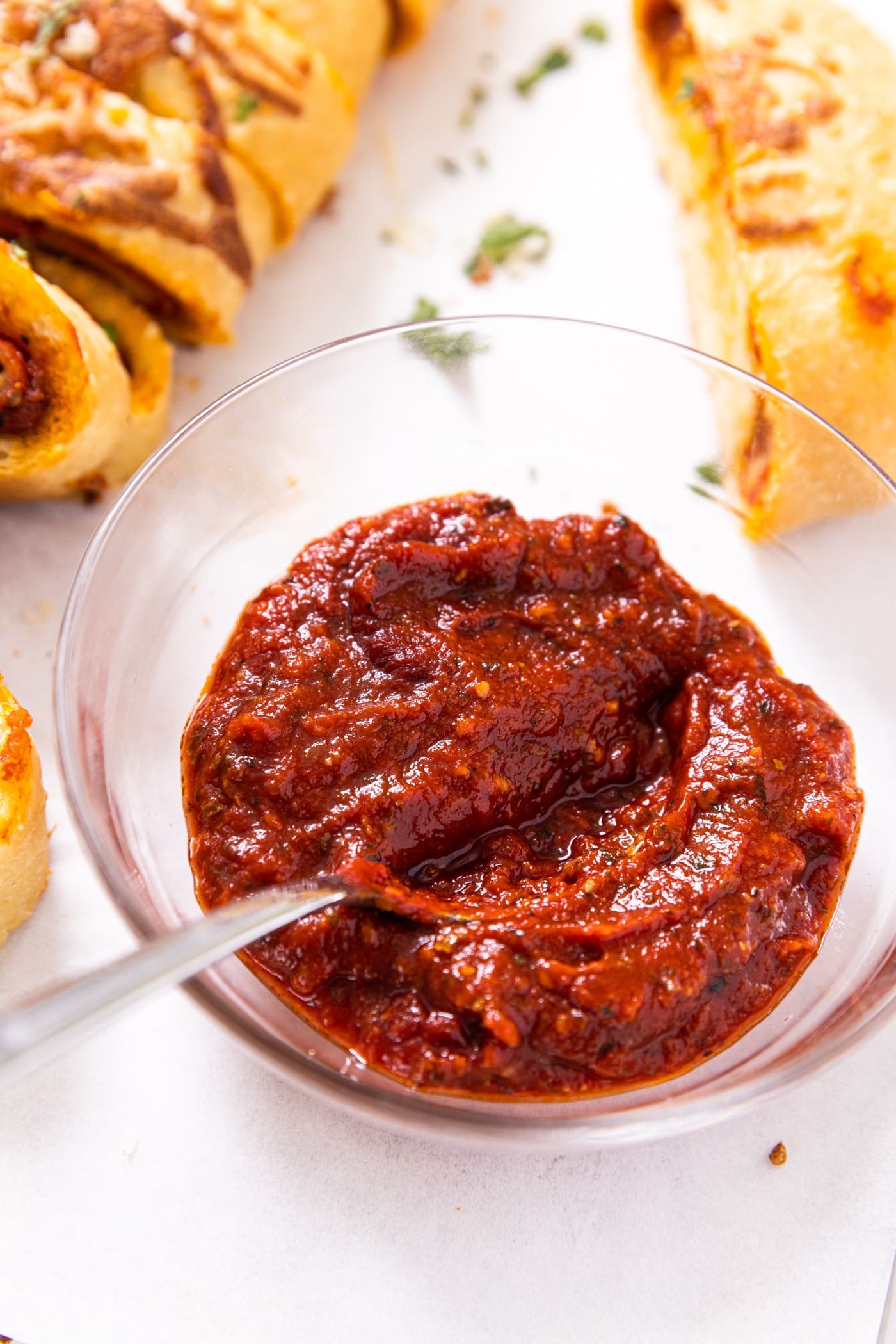 Small glass bowl filled with red tomato sauce with spoon in sauce sitting on white piece of parchment paper with slices of stromboli beside
