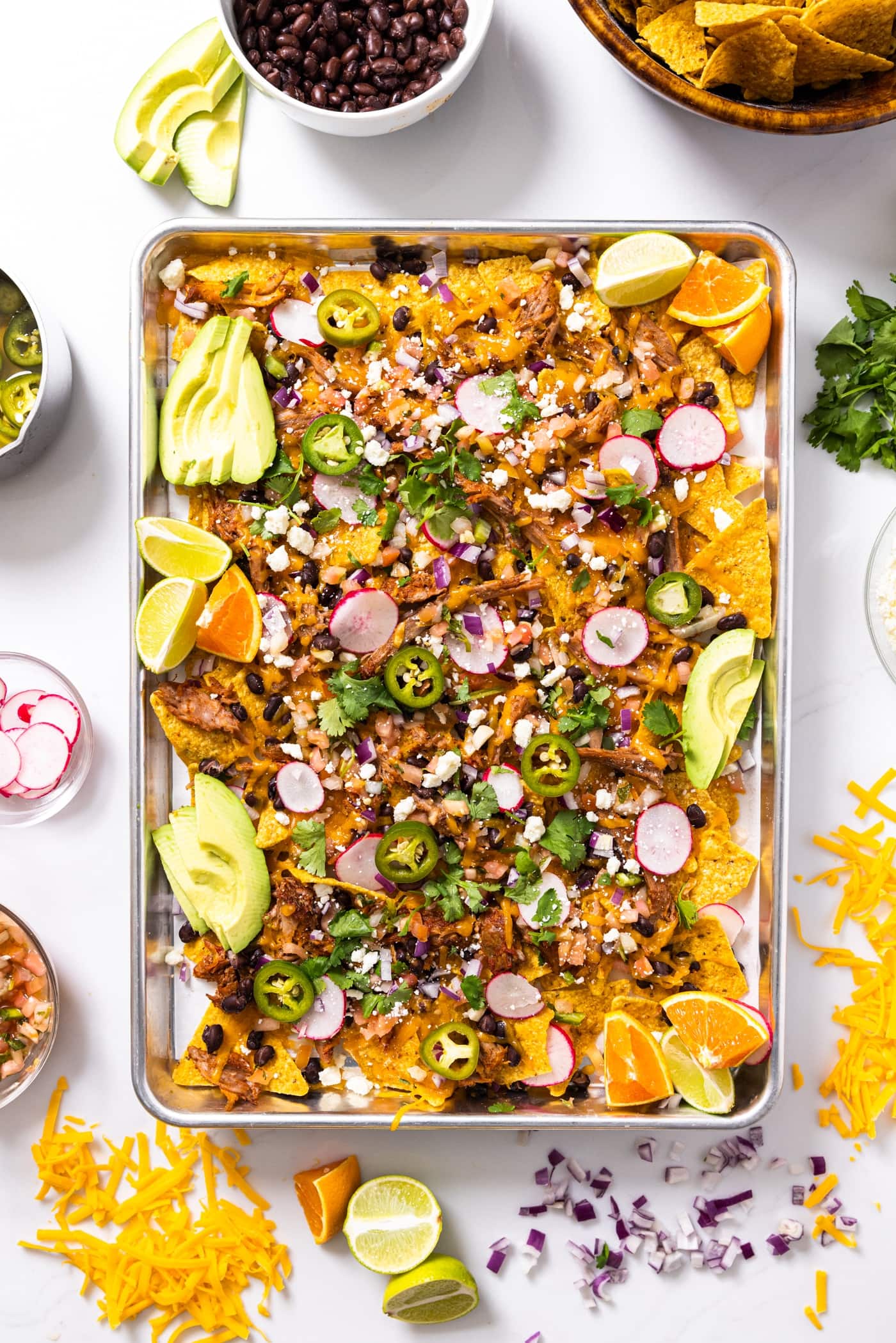 Top down view of metal sheet pan sitting on white countertop filled with tortilla chips and pulled pork with lots of toppings on top along with shredded cheese