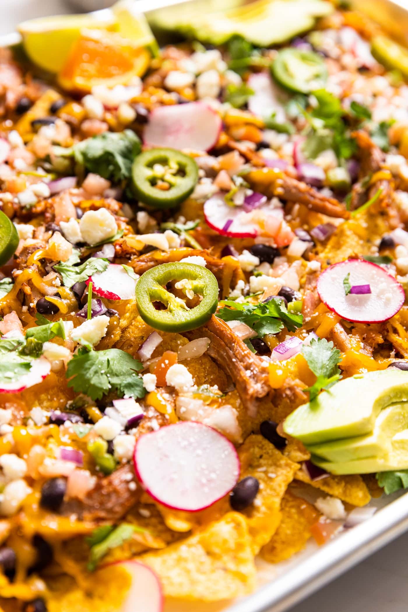Large metal sheet pan with lots of tortilla chips underneath topped with pulled pork and lots of toppings and shredded cheese