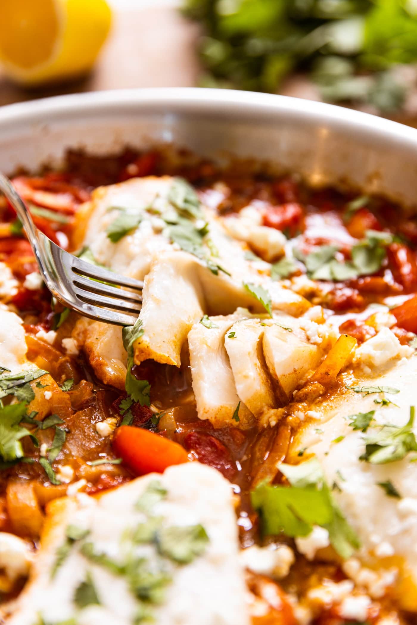 Fork holding piece of filet of cod sitting amongst red tomato sauce topped with feta and chopped cilantro with lemon in background