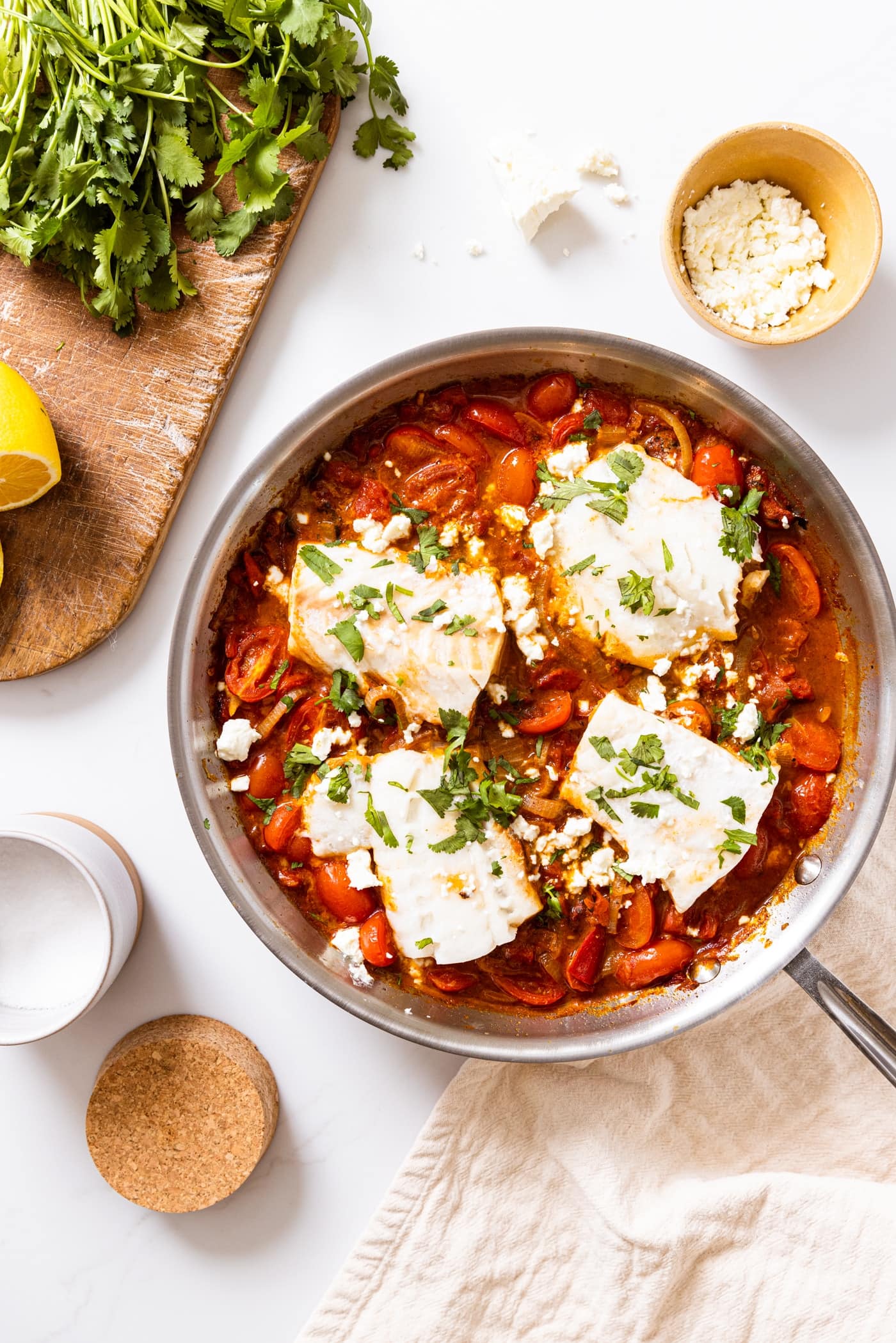 Top down view of white countertop with metal skillet filled with red tomato sauce with filets of cod nestled inn with lemon and cilantro sitting on cutting board beside skillet