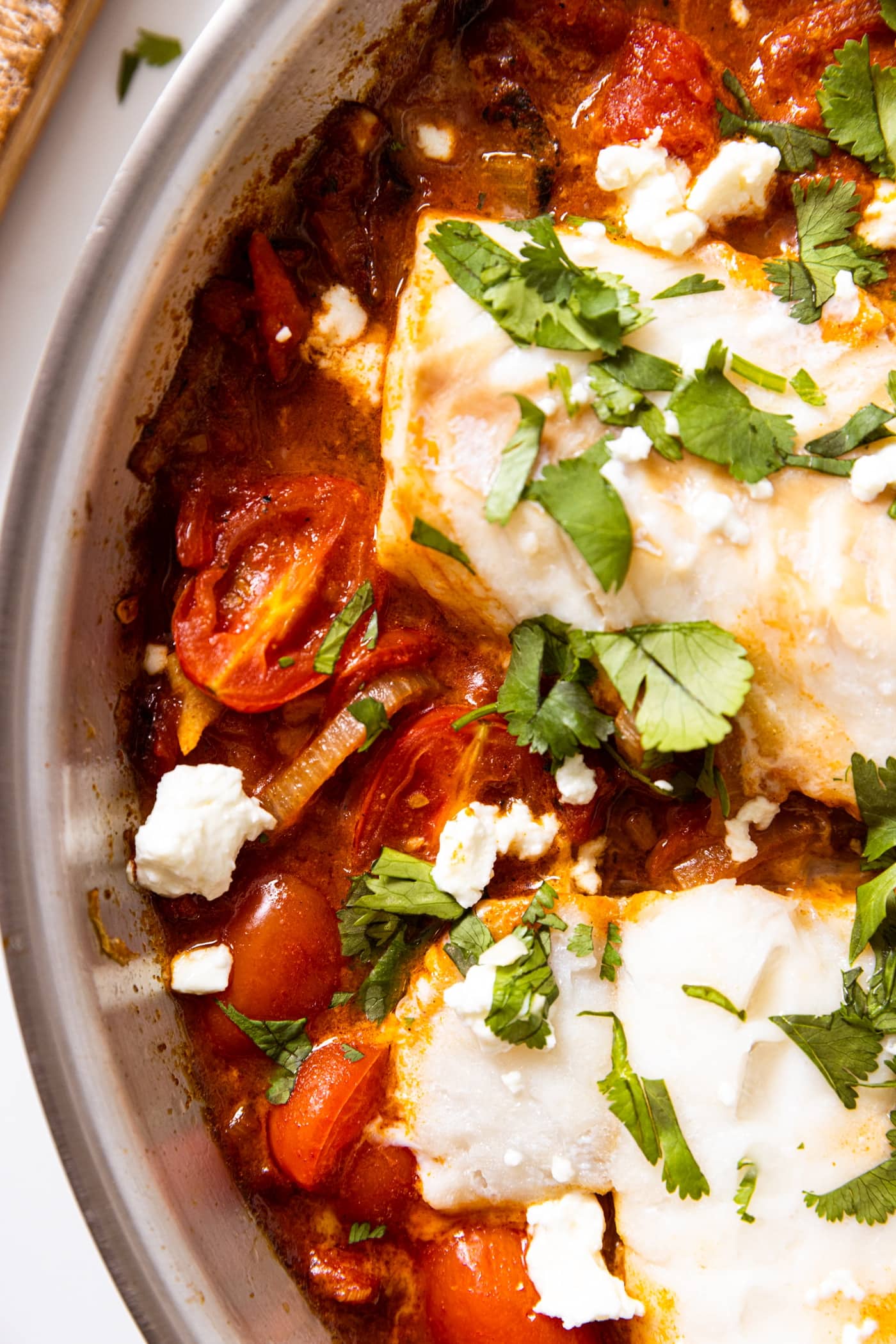 Top down view of red tomato sauce with half of grape tomato showing with filets of cod sitting around topped with cilantro and feta all on white countertop