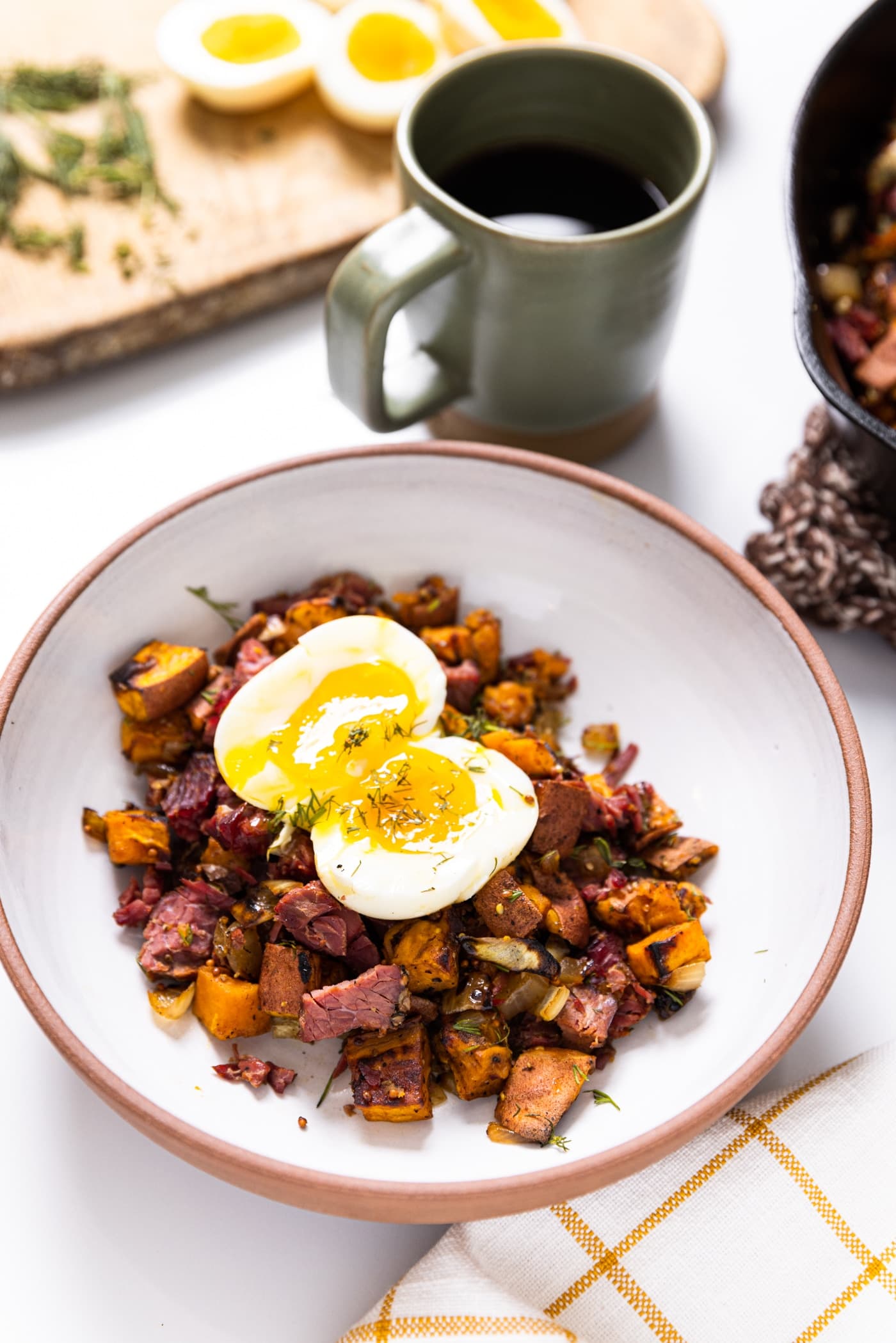 White ceramic bowl filled with sweet potato hash along with corned beef topped with a jammy egg sitting on white countertop with green mug filled with coffee in background