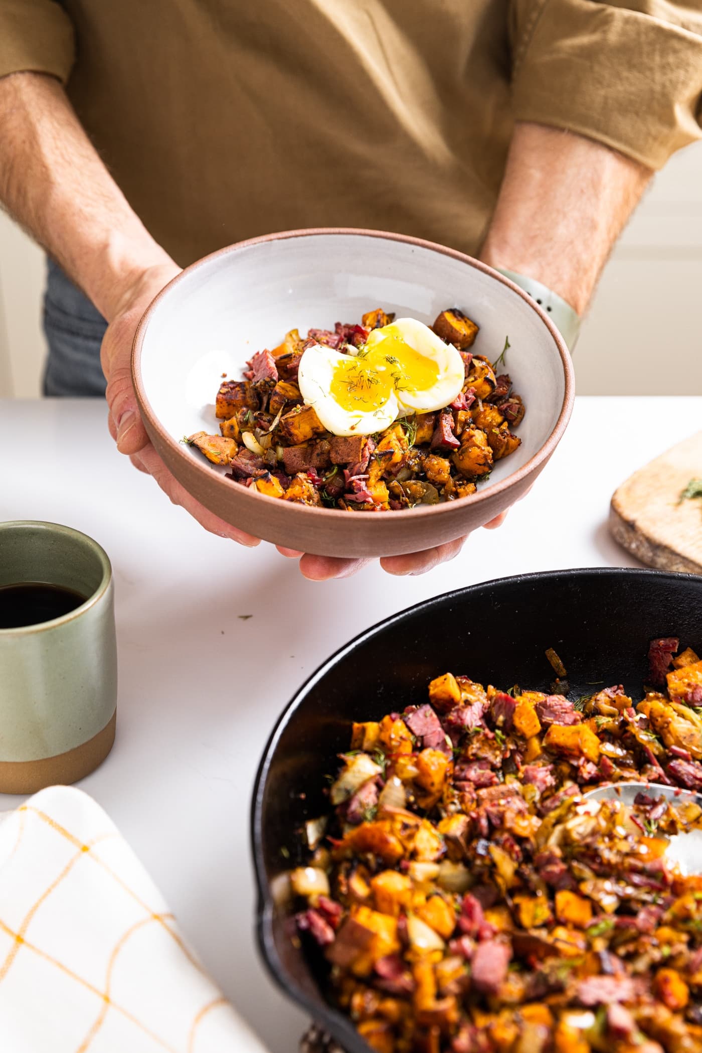Two hands holding white and brown ceramic bowl filled with corned beef and sweet potato hash with jammy egg on top with skillet and rest of hash sitting below
