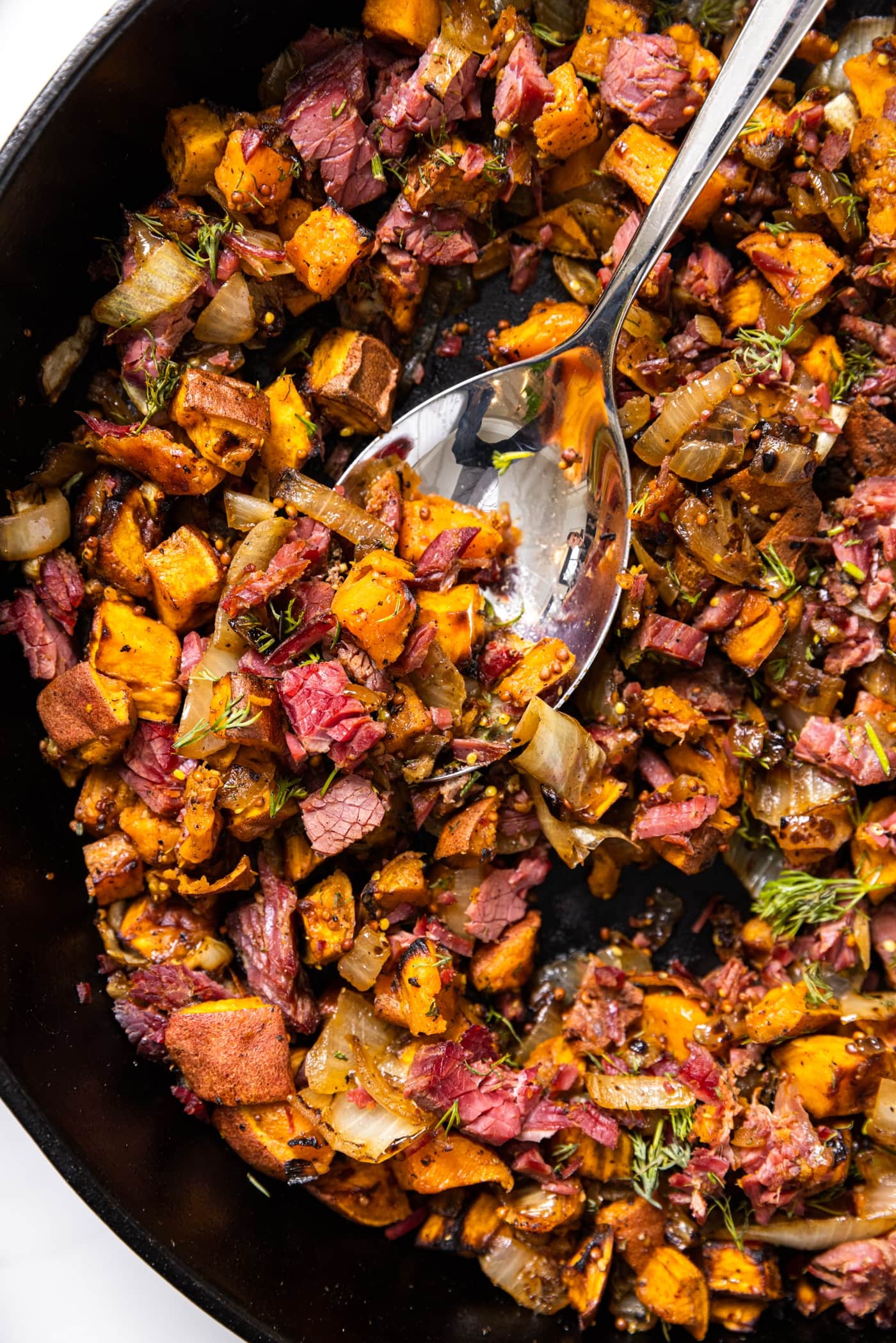 Top down view of sweet potato hash mixed with corned beef sitting in black skillet with spoon taking a scoop
