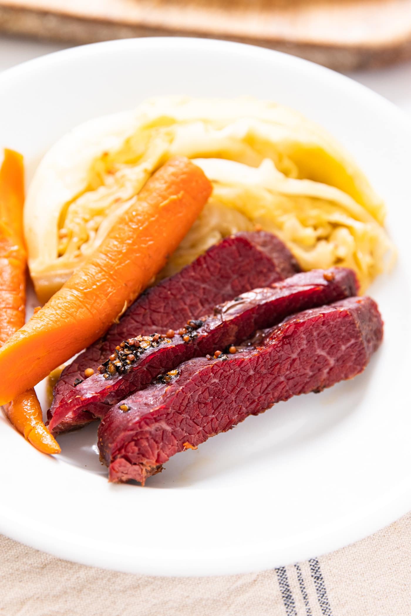 Close up view of three slices of red colored corned beef sitting on white plate with cabbage and carrots in background