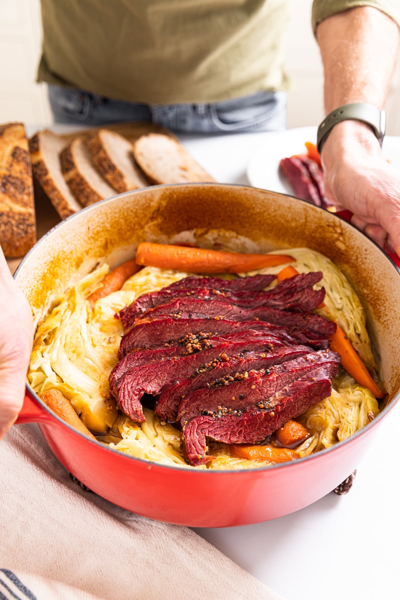 Large red Dutch oven with two hands holding handles filled with red colored corned beef sitting on top of cabbage and carrots on white countertop