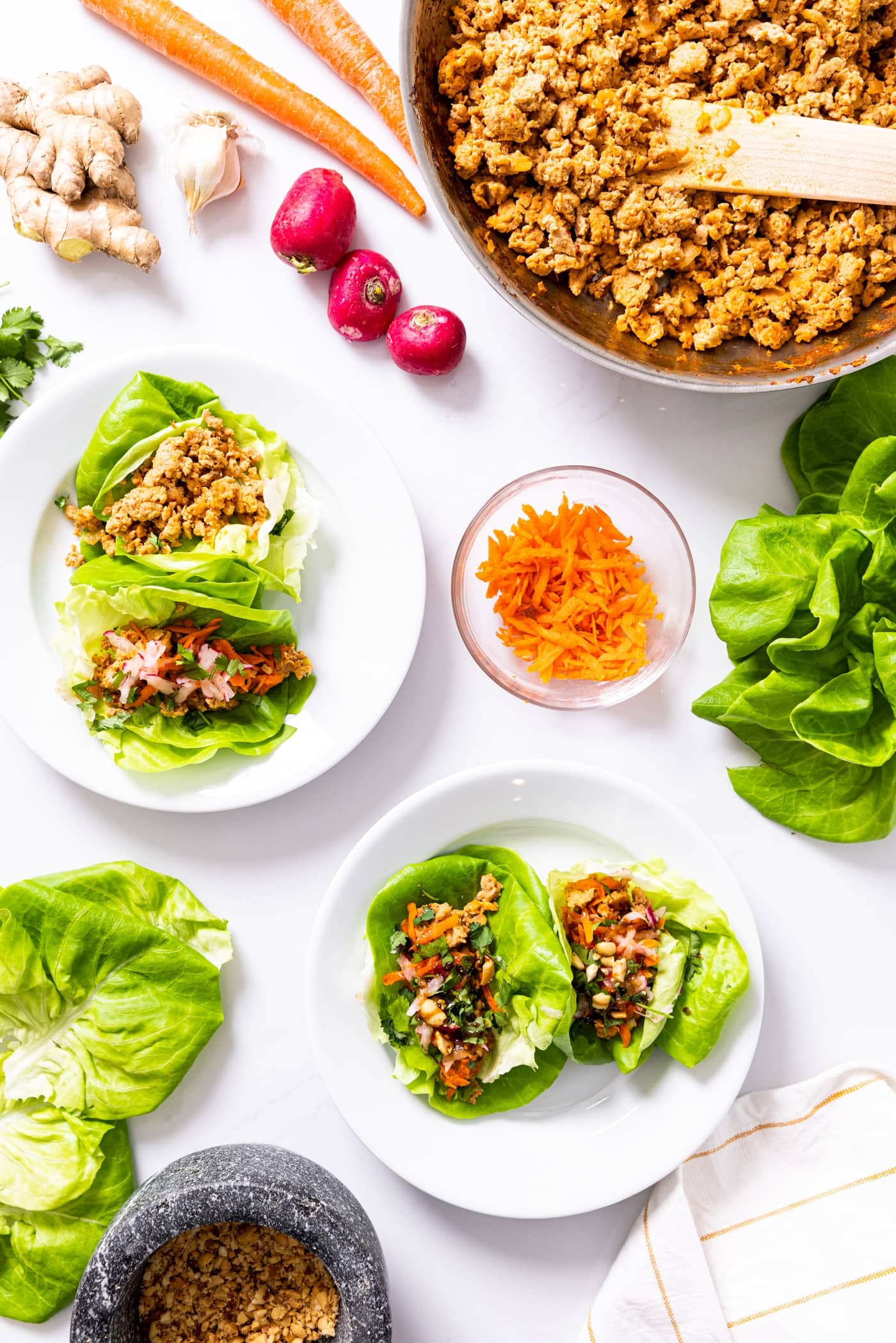 Top down view of white countertop with white plates filled with chicken lettuce cups with carrots and radishes as toppings with skillet with rest of chicken