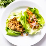 Top down view of white plate filled with two chicken lettuce cups one with radish and carrots as toppings all on white countertop