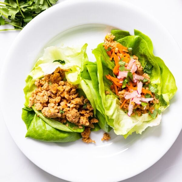 Top down view of white plate filled with two chicken lettuce cups one with radish and carrots as toppings all on white countertop