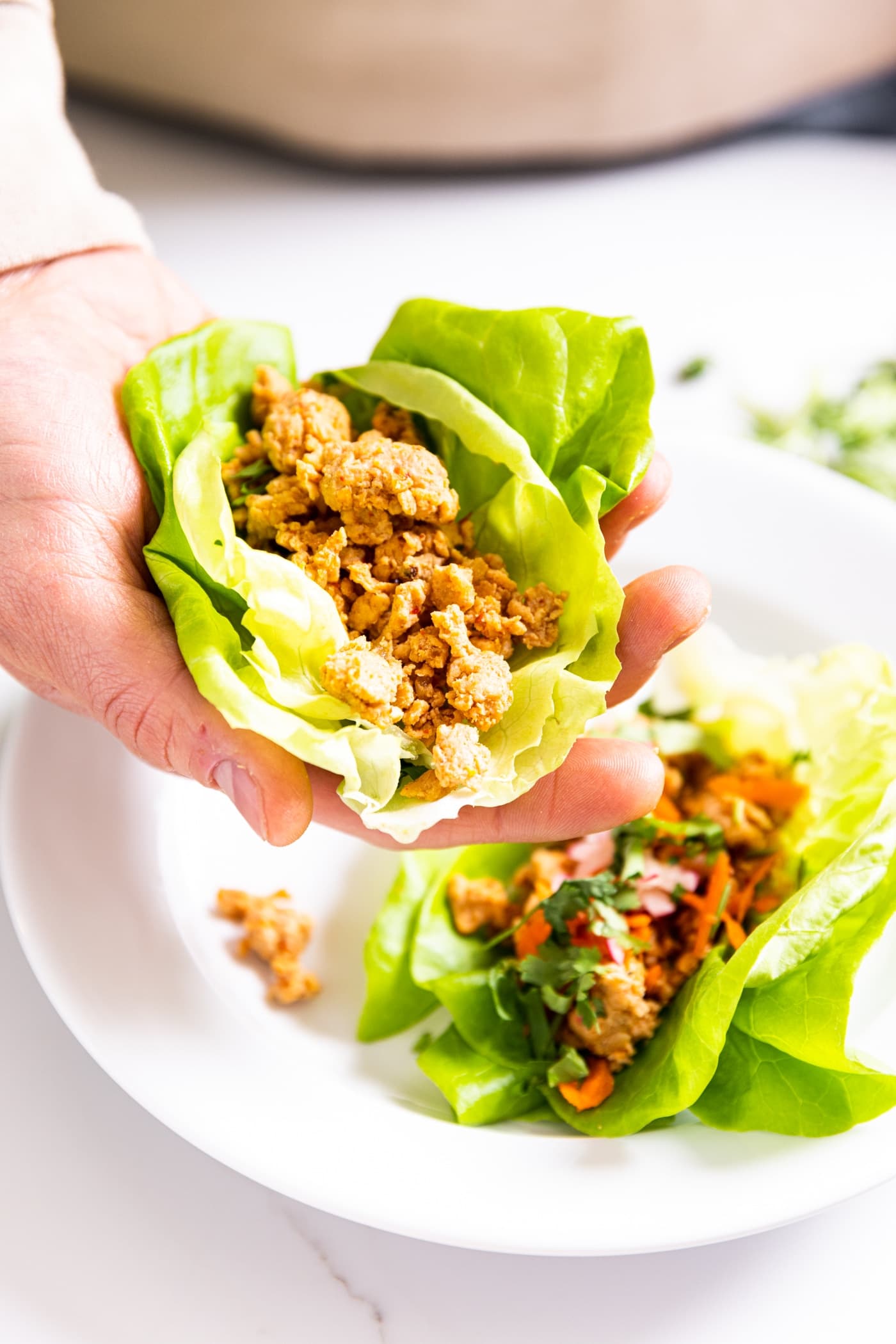 Hand holding green lettuce cup filled with spicy chicken with another lettuce cup sitting on white plate in background