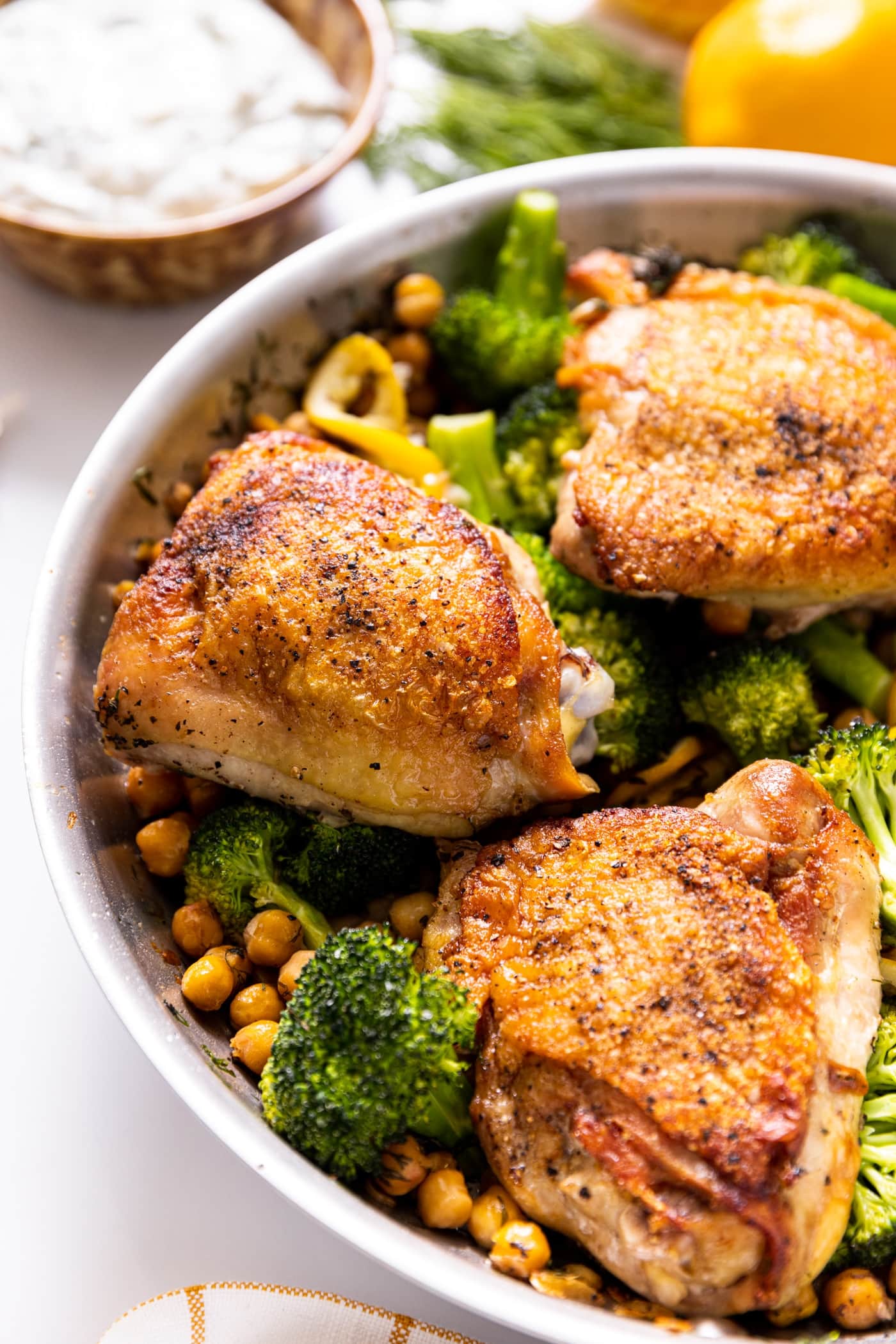Metal skillet sitting on white countertop with three chicken thighs with crispy skin sitting on top of broccoli and chickpeas