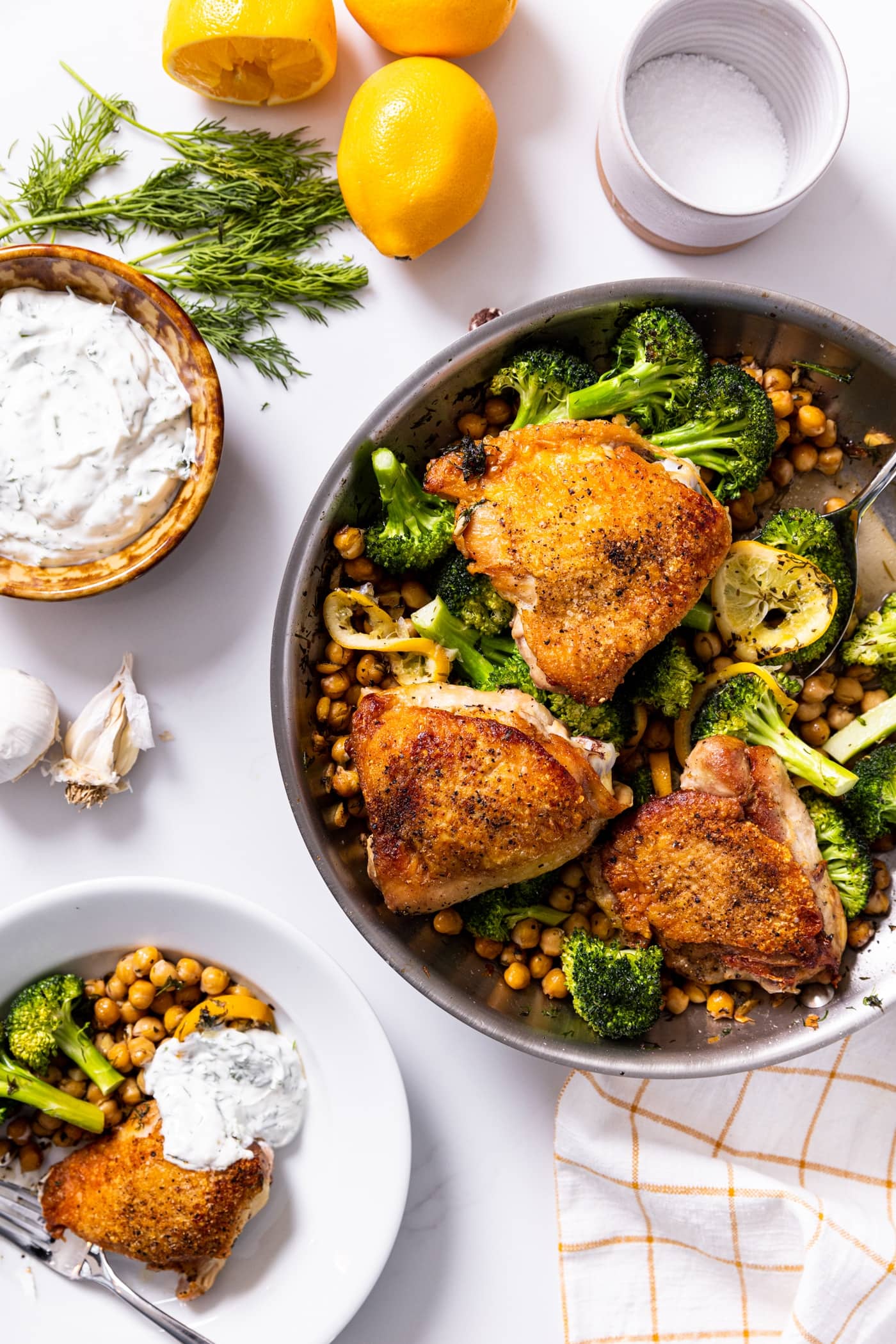 Top down view of white countertop with metal skillet filled with chicken thighs, broccoli, and chickpeas
