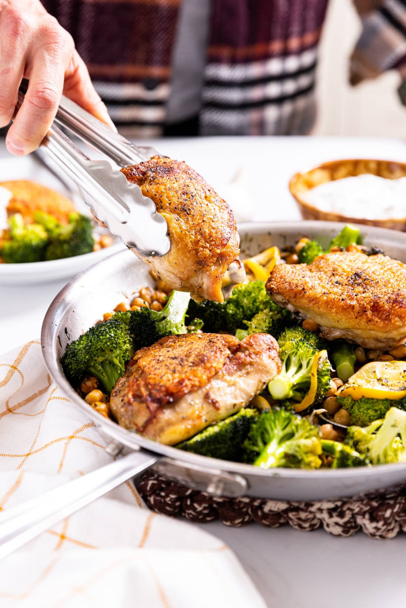 Hand holding tongs lifting up a chicken thigh from a metal skillet with broccoli and chickpeas