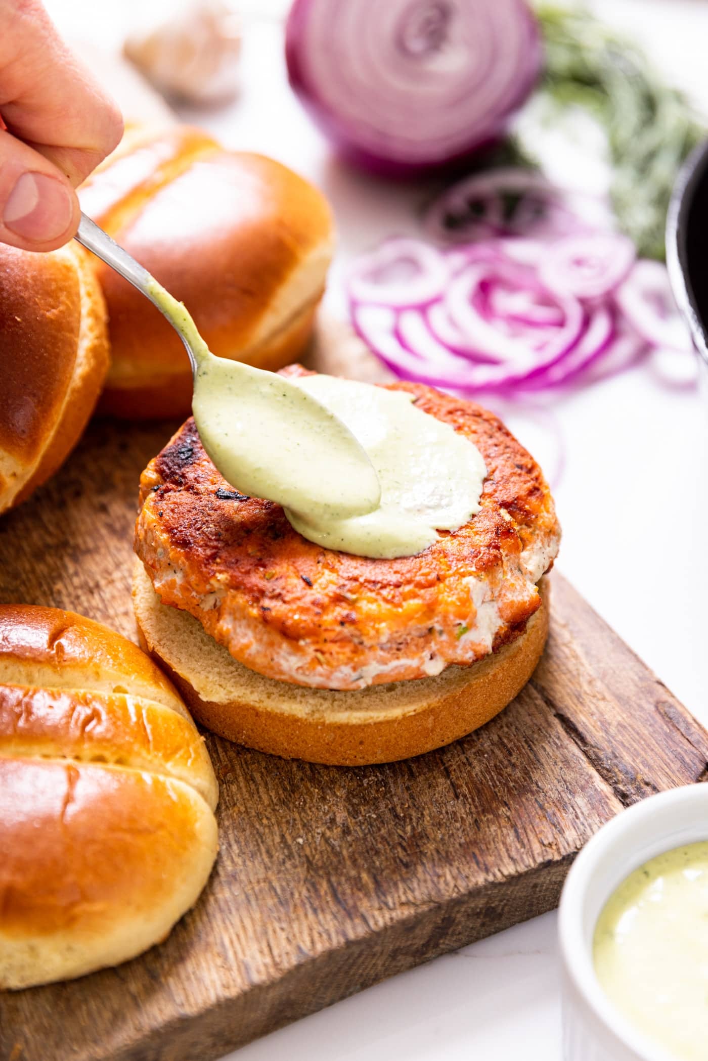 Hand holding spoon smoothing dill aioli over the top of salmon burger sitting on brioche bun sitting on wood cutting board