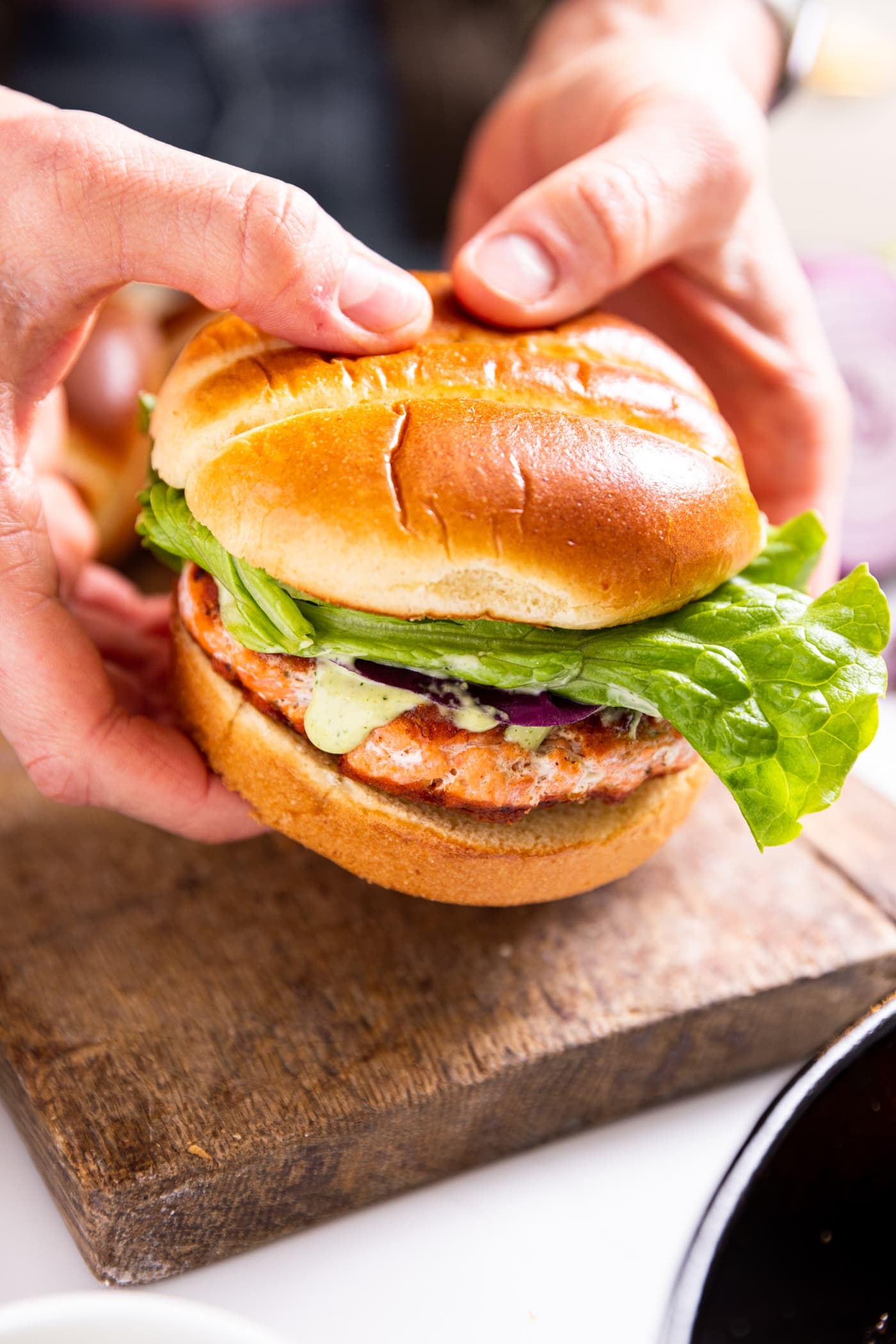 Two hands holding salmon burger in brioche buns with piece of lettuce and dill aioli inside with wood board underneath