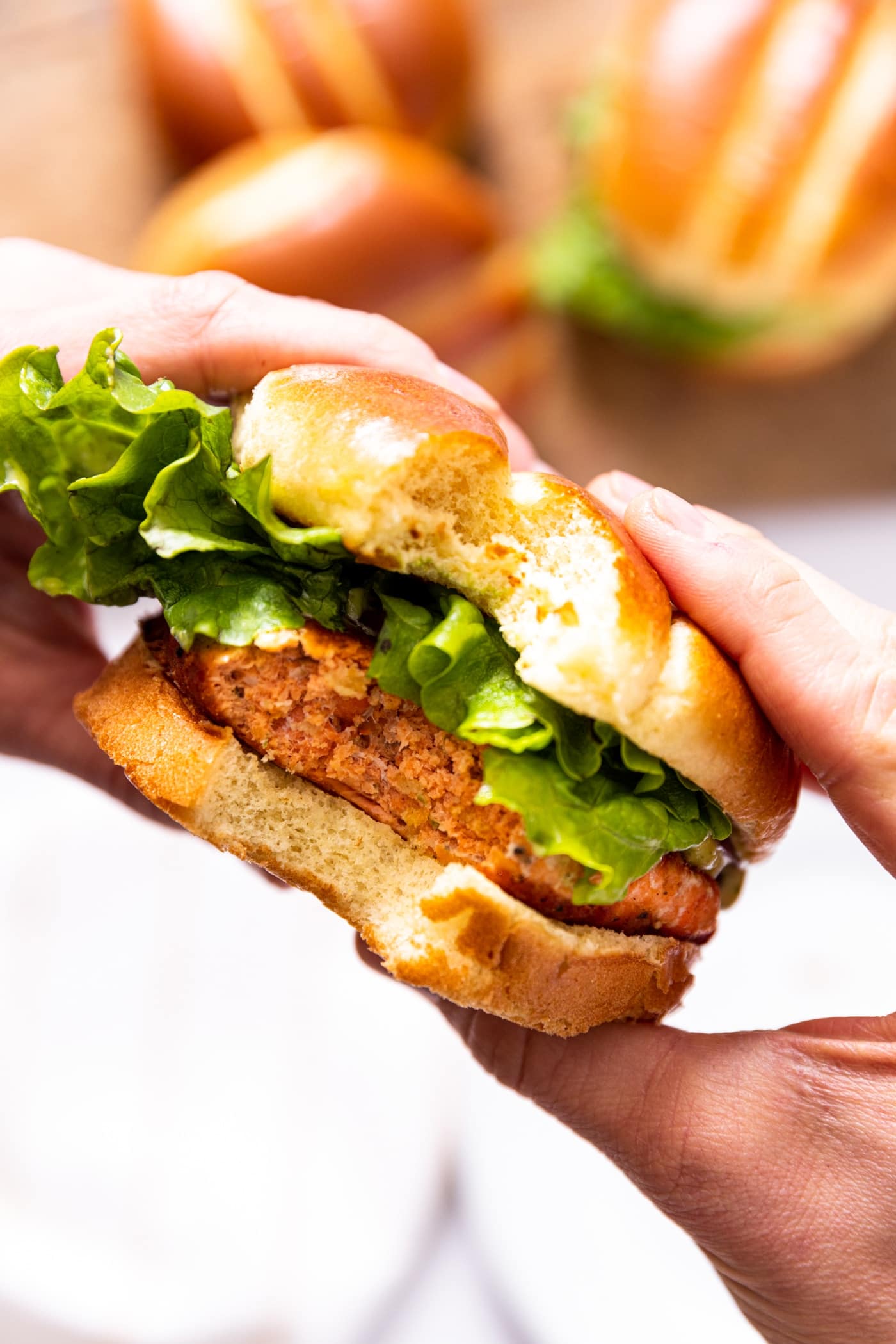 Two hands holding salmon burger with piece of lettuce on top with brioche bun with two other burgers in background