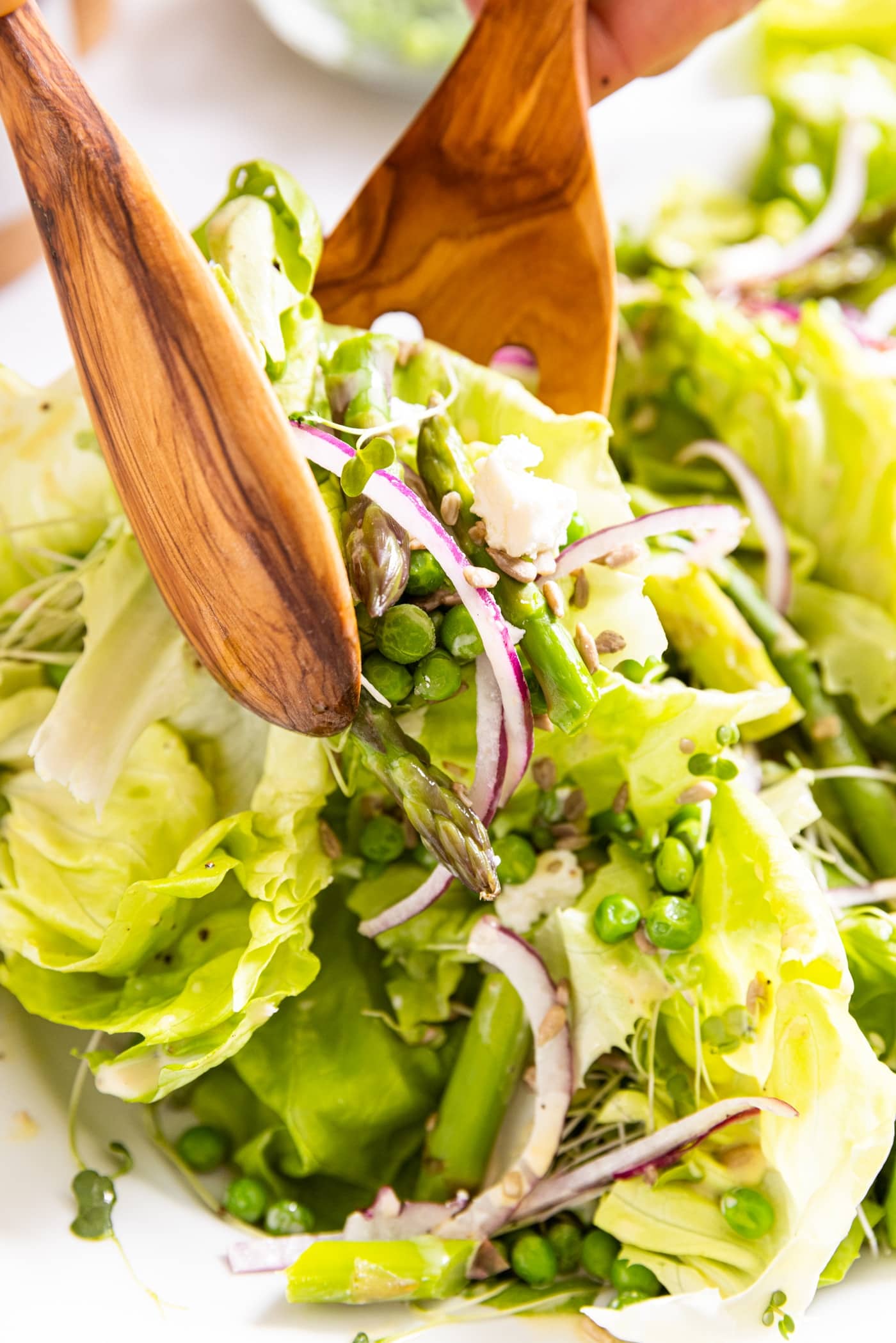 Two wooden tongs holding serving of salad made of buttercrunch lettuce and red onion and peas with white serving platter underneath