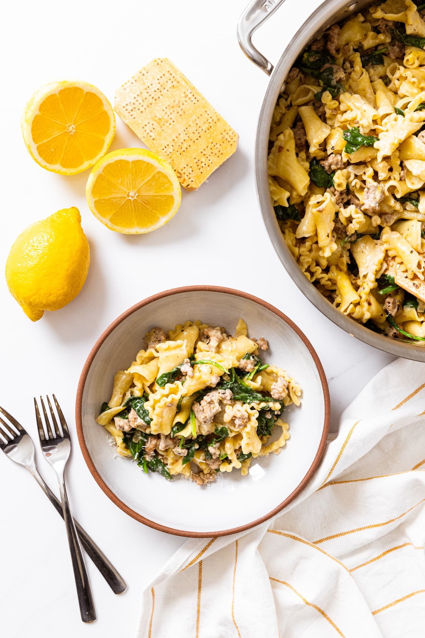 Top down view of white countertop with sliced lemons and bowl filled with yellow colored pasta with spinach and sausage and forks to the side