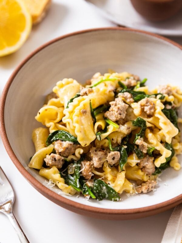 Ceramic bowl with brown rim sitting on white countertop with heaping pile of yellow colored pasta with bits of sausage and spinach with lemon in the background
