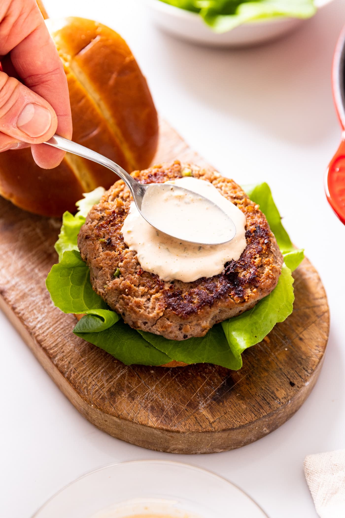 Hand holding spoon spreading taupe colored sauce on top of turkey burger with slice of lettuce on top of brioche bun all on white countertop