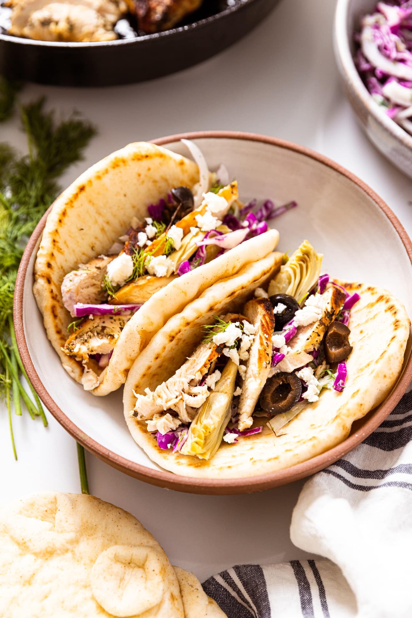 Ceramic bowl holding two pitas stuffed with grilled chicken and cabbage slaw with skillet in background as well as cabbage slaw container