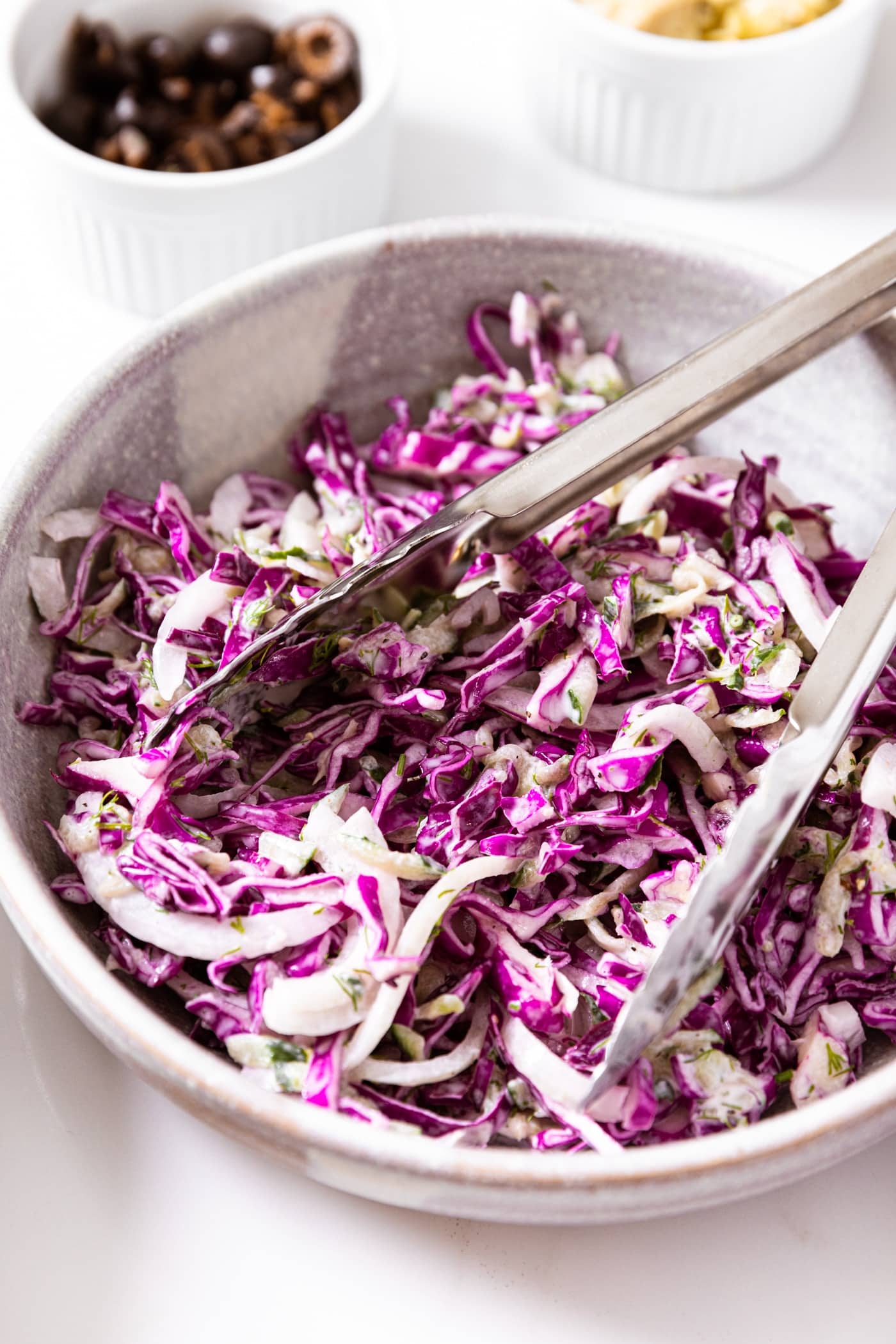 White bowl filled with red cabbage topped with creamy Greek yogurt dressing with metal tongs in middle of bowl