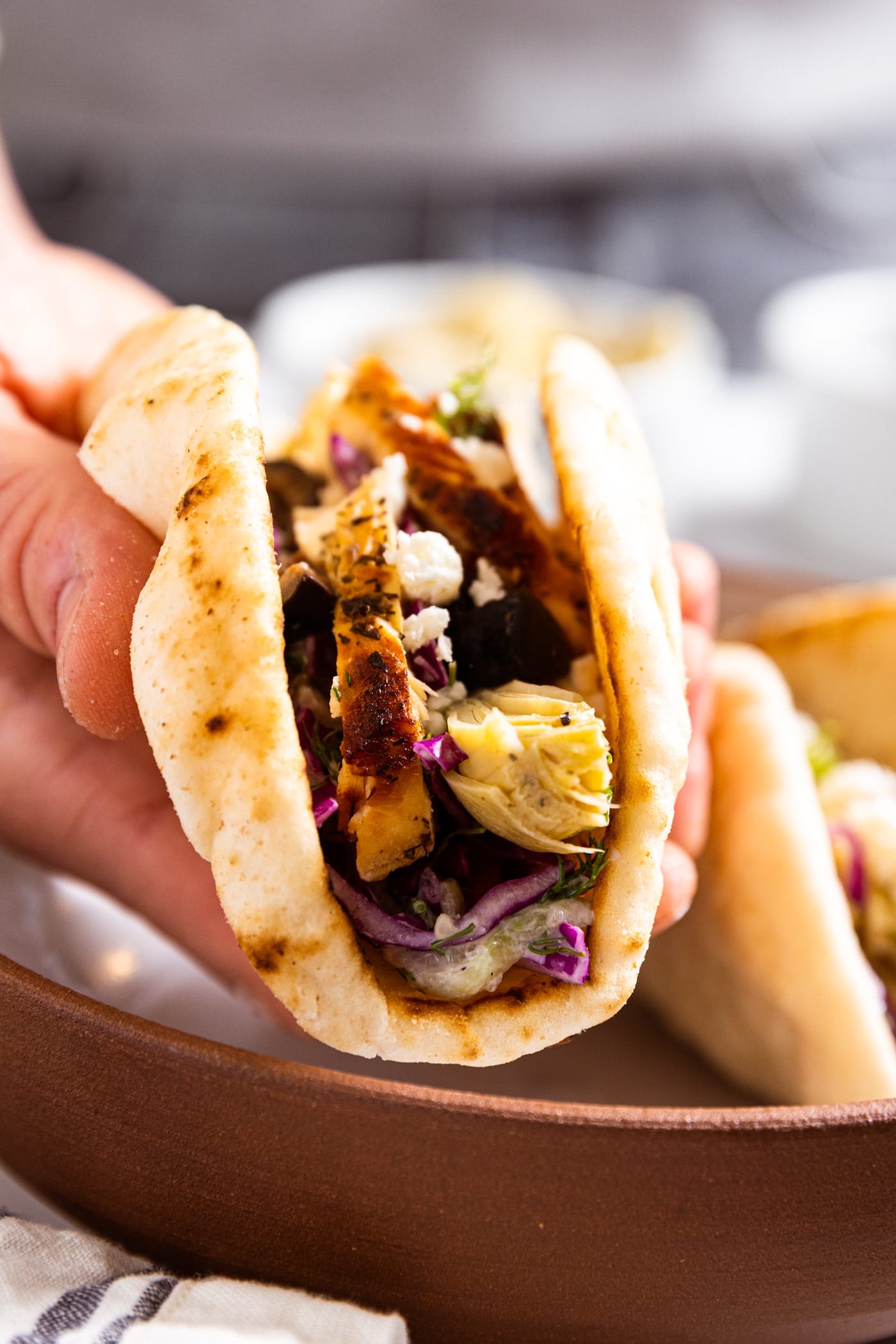 Hand holding pita shell with grilled chicken and artichoke hearts inside with plate in background