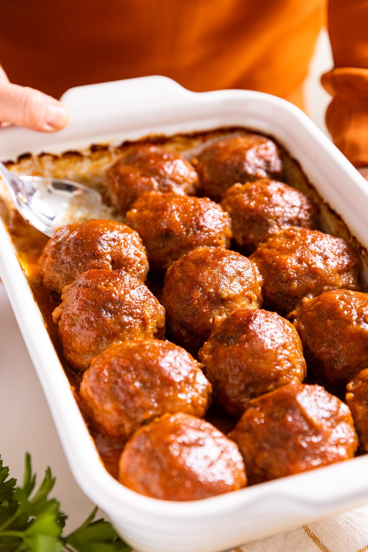 Hand holding white baking dish filled with brown colored ham balls with sauce on top with silver spoon sitting in dish
