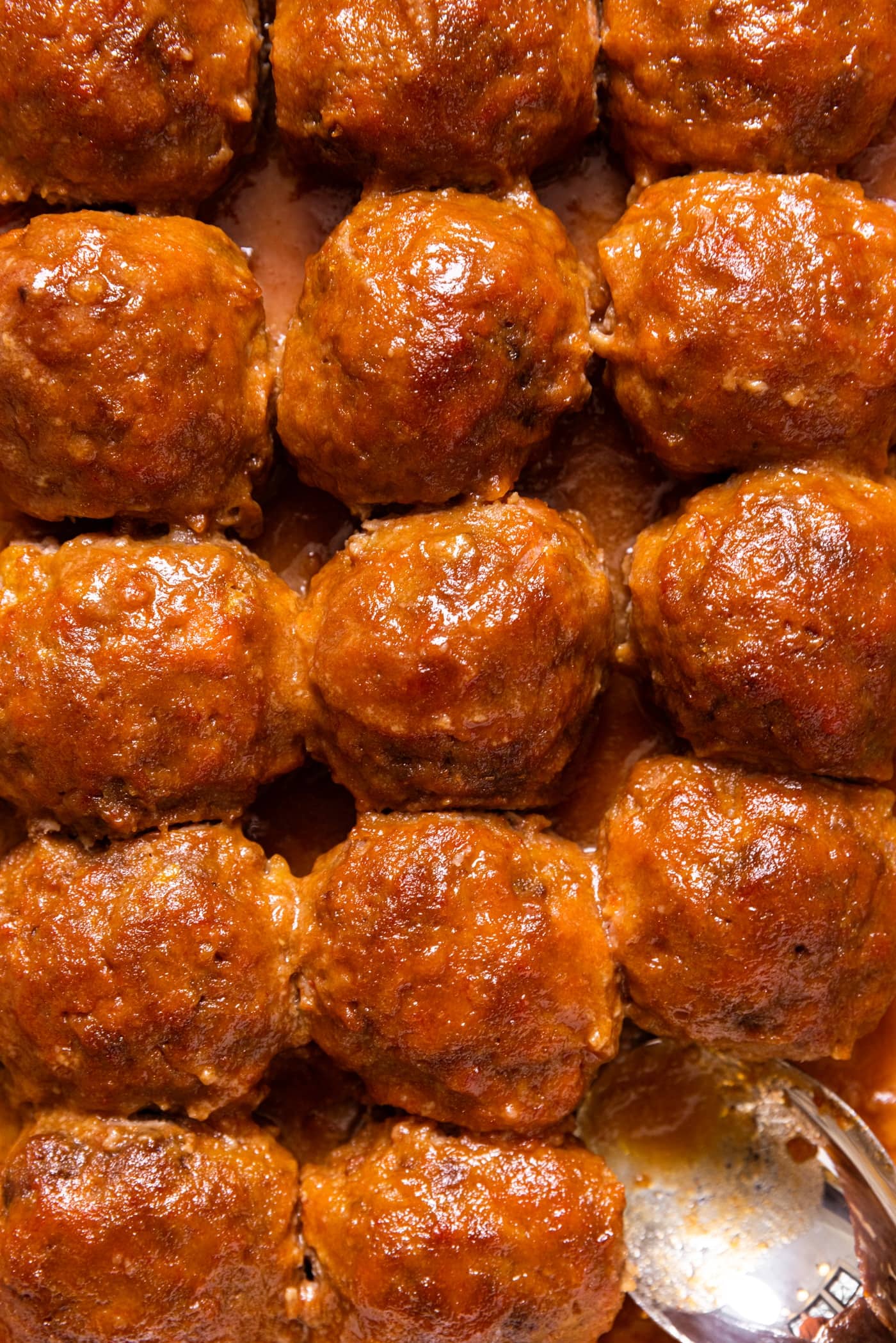 Top down view of brown colored ham balls with sauce all over the top sitting in white baking dish with spoon